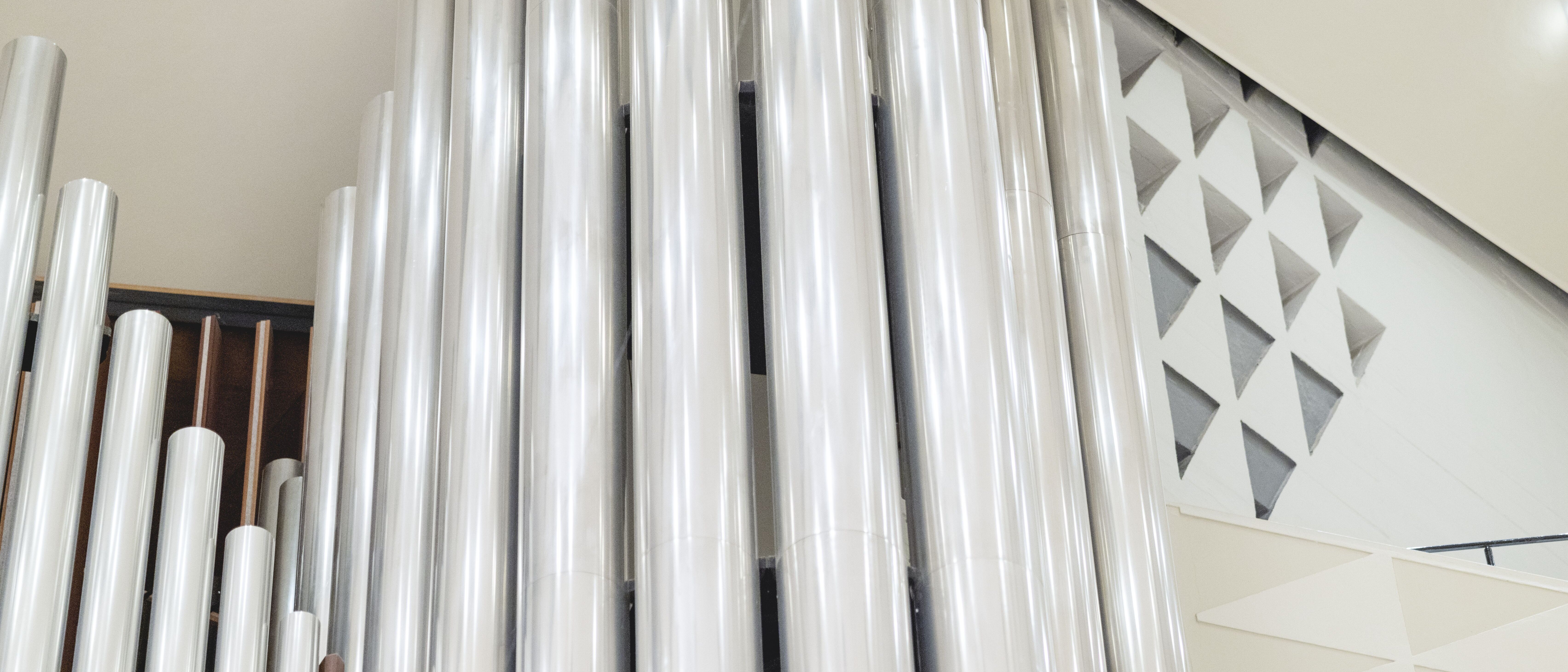 Große, silbern glänzende Orgelpfeifen stehen vertikal im Raum, mit geometrischen Wandmustern und heller Beleuchtung im Hintergrund, was eine moderne und klare architektonische Szene schafft.