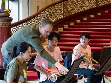 Posaunist Jesper Busk Sørensen beugt sich vor und zeigt auf Notenblätter, während er drei junge Musiker mit Blechblasinstrumenten anleitet, die vor Notenständern in der Nähe einer großen Treppe sitzen.