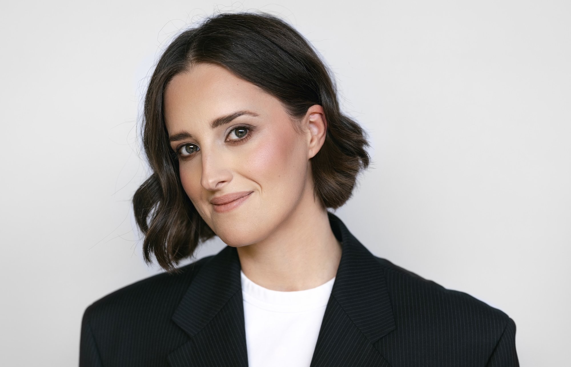 A woman with short brown hair, wearing a black oversized blazer and white shirt, smiles softly at the camera. She holds rings in her hands against a plain light background.