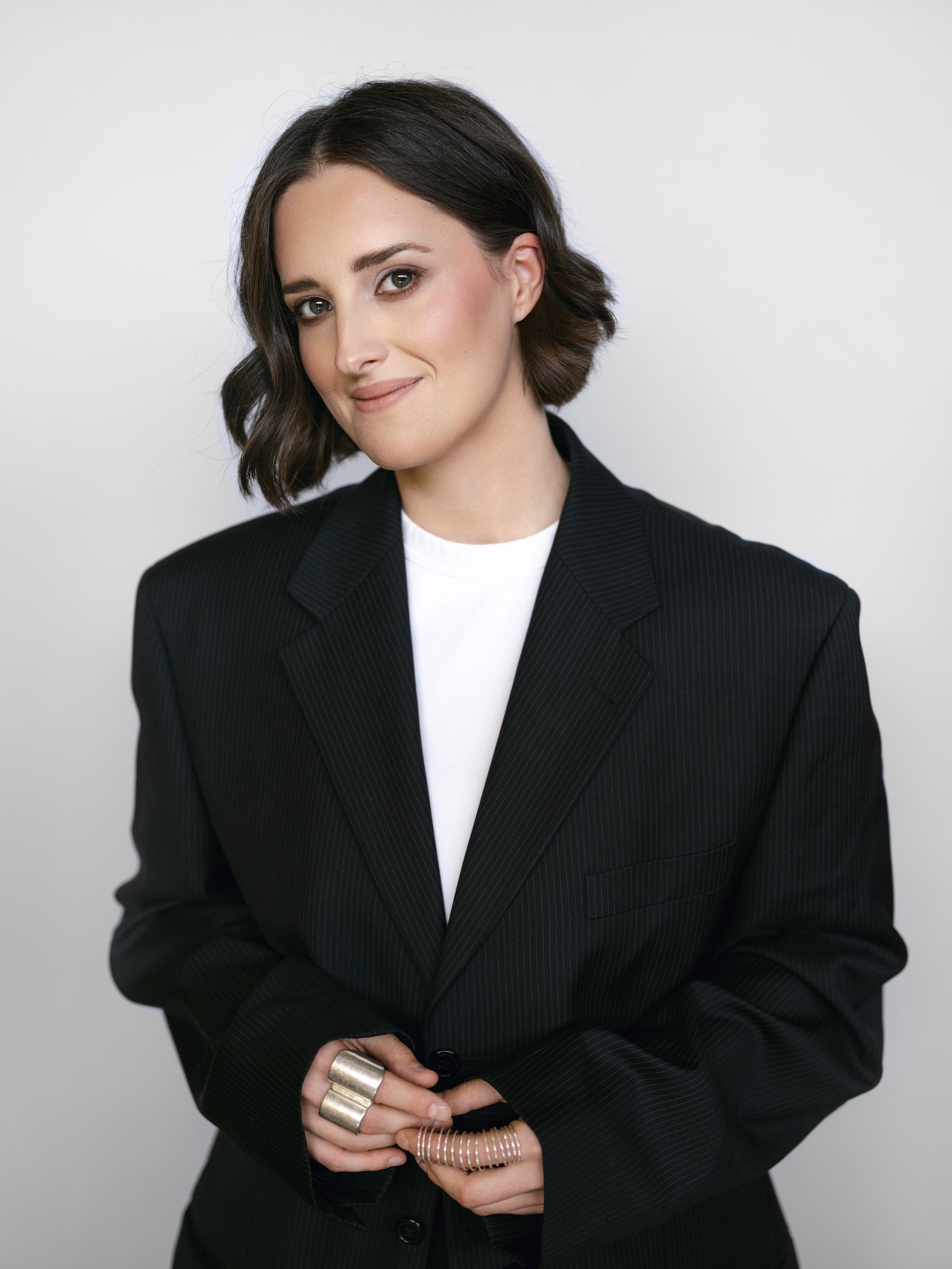 Eine Frau mit kurzen, dunklen Haaren trägt einen schwarzen Blazer über einem weißen Hemd, lächelt leicht, hat die Hände gefaltet und Ringe an den Fingern und steht vor einem schlichten, hellen Hintergrund.