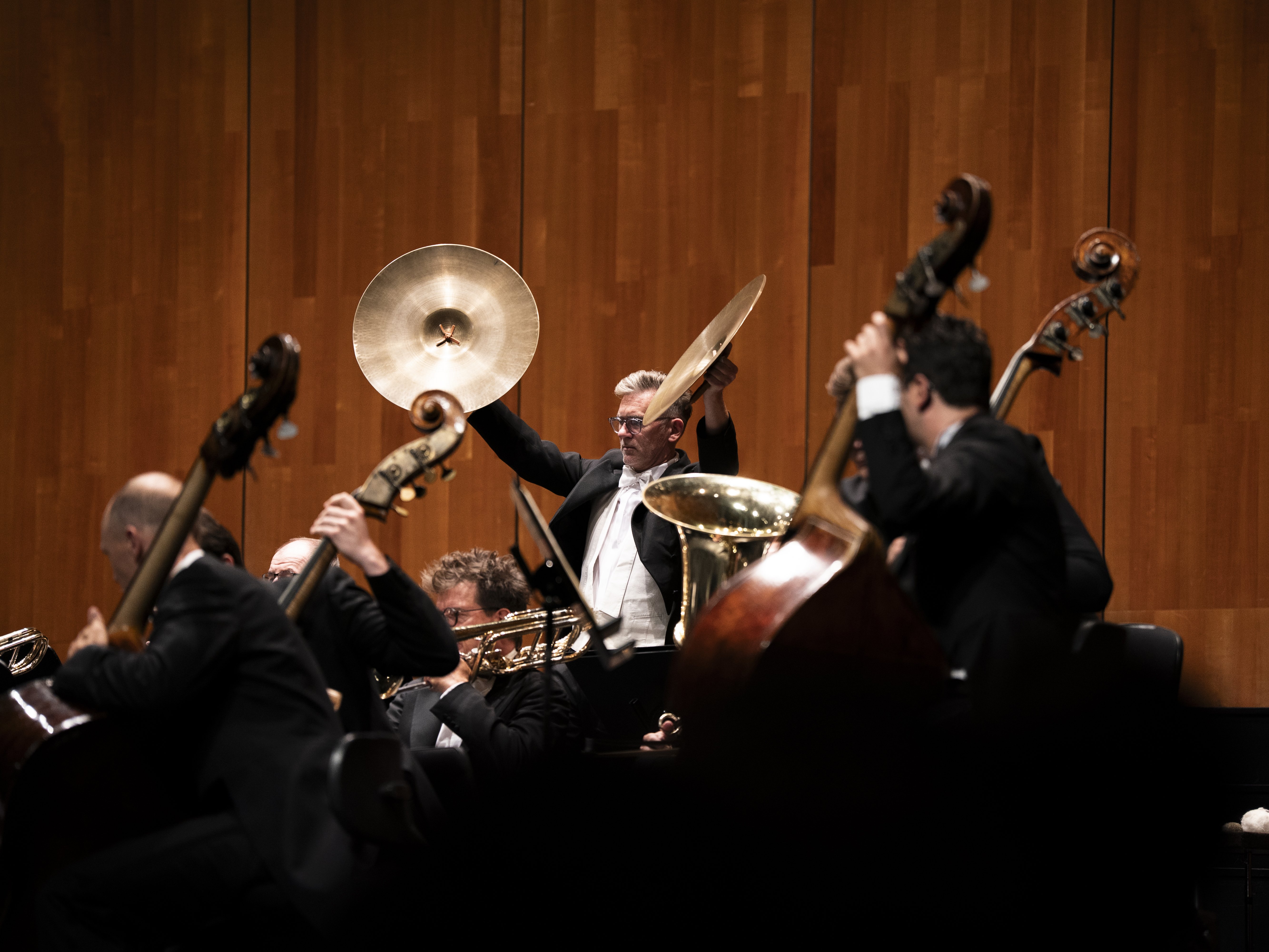 Ein Musiker hebt während eines Auftritts mit einem Orchester auf der Bühne die Becken. Andere Musiker spielen Blech- und Streichinstrumente. Im Hintergrund ist eine Holzwand zu sehen.