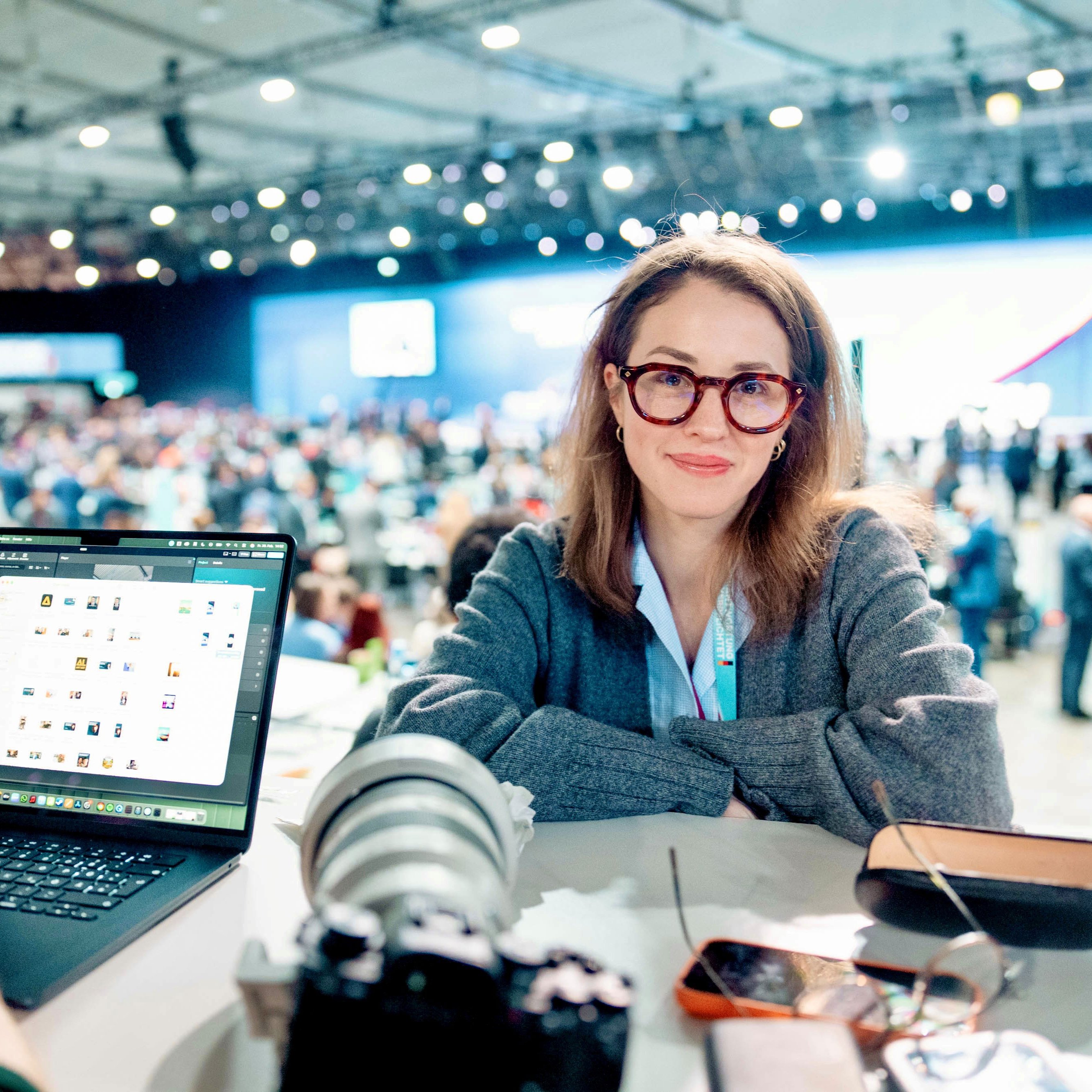 Eine Frau mit Brille sitzt an einem Schreibtisch, auf dem ein Laptop, eine Kamera, ein Handy und eine Wasserflasche stehen. Im Hintergrund des hell beleuchteten Raums sind eine große, verschwommene Menschenmenge und eine Bühne zu sehen.