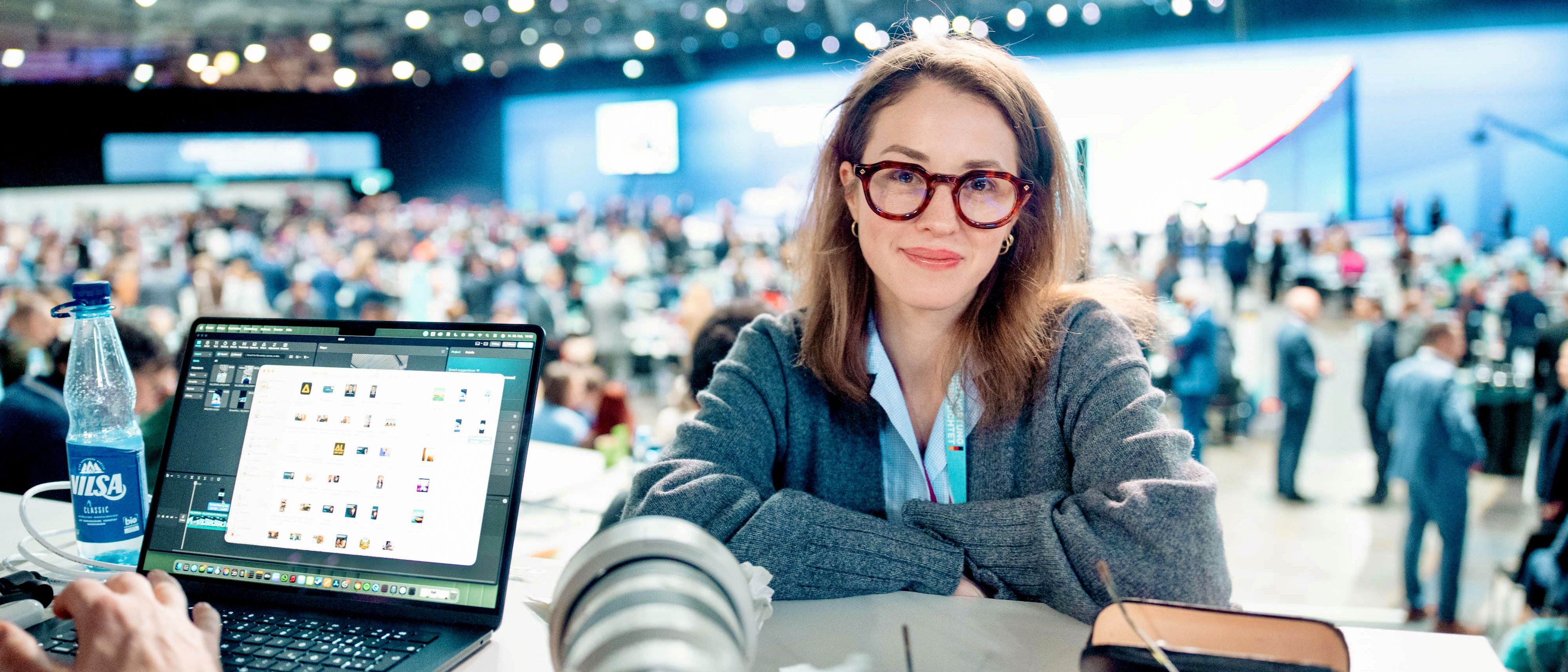 Eine Frau mit Brille sitzt an einem Schreibtisch, auf dem ein Laptop, eine Kamera, ein Handy und eine Wasserflasche stehen. Im Hintergrund des hell beleuchteten Raums sind eine große, verschwommene Menschenmenge und eine Bühne zu sehen.