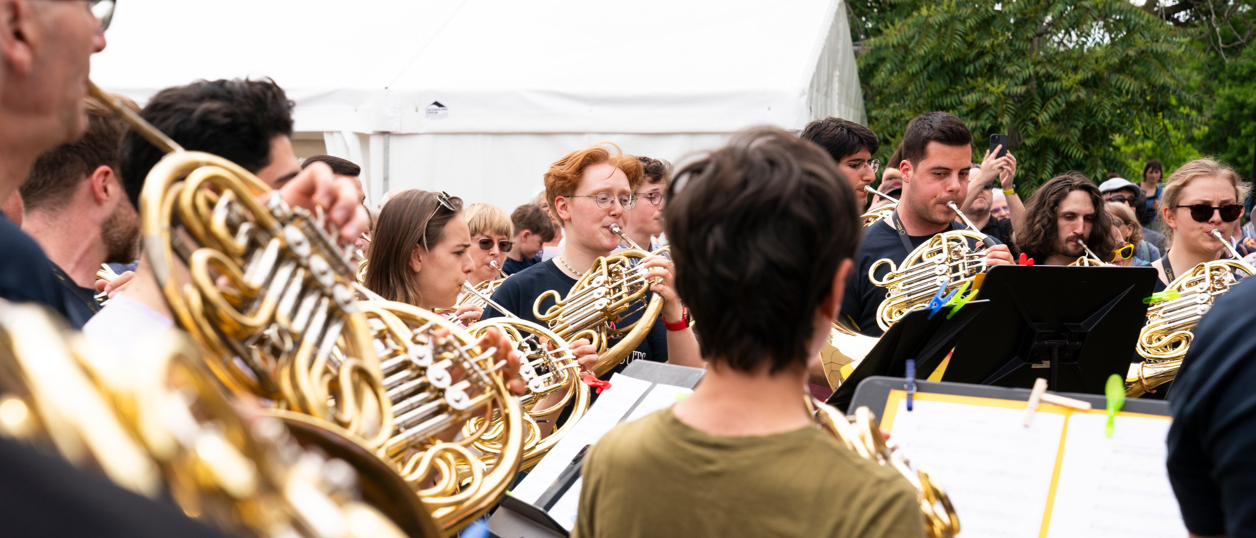 Eine große Gruppe von Menschen im Freien, die gemeinsam Waldhorn spielen. Sie stehen dicht beieinander und lesen Noten, im Hintergrund sind Bäume und ein weißes Zelt zu sehen.