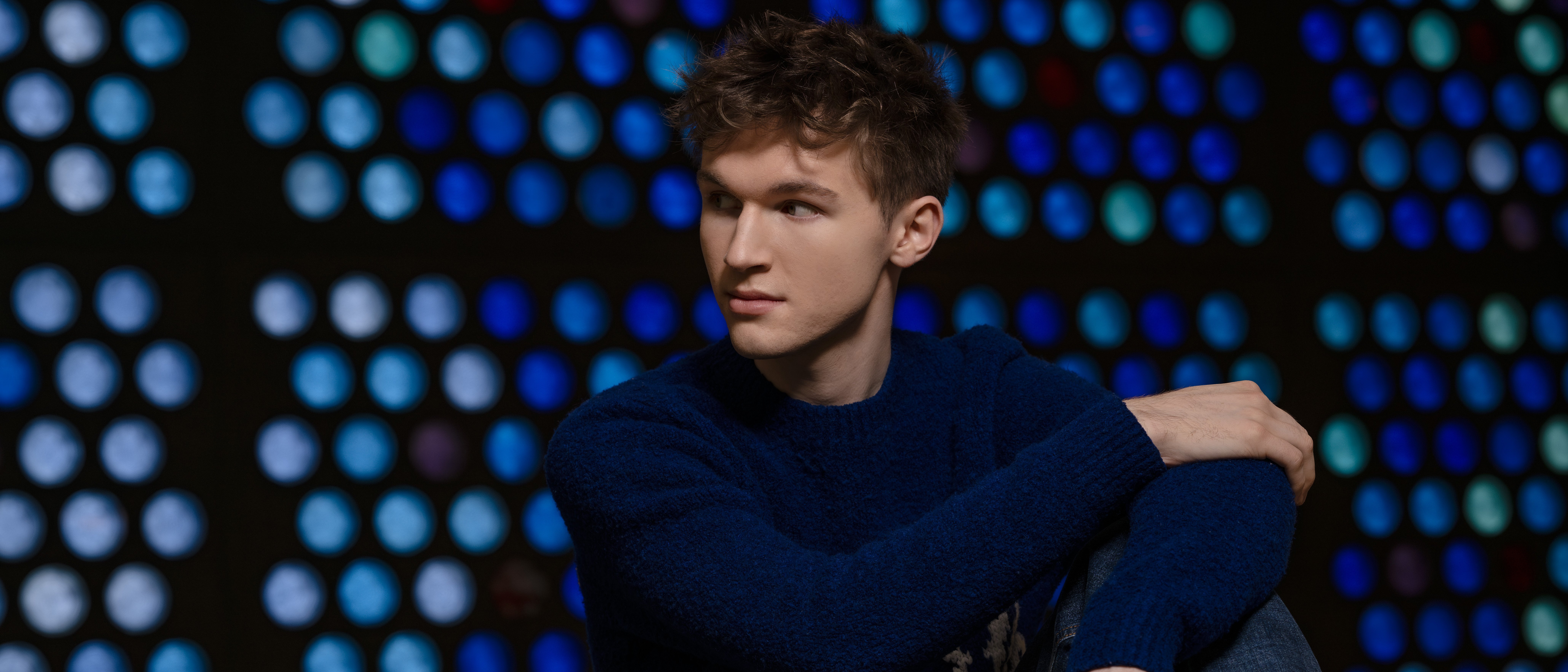 Portrait of the young man Jan Liebermann, sitting in a blue jumper and jeans in front of a background with blue, round glass windows. He is looking to the side. 