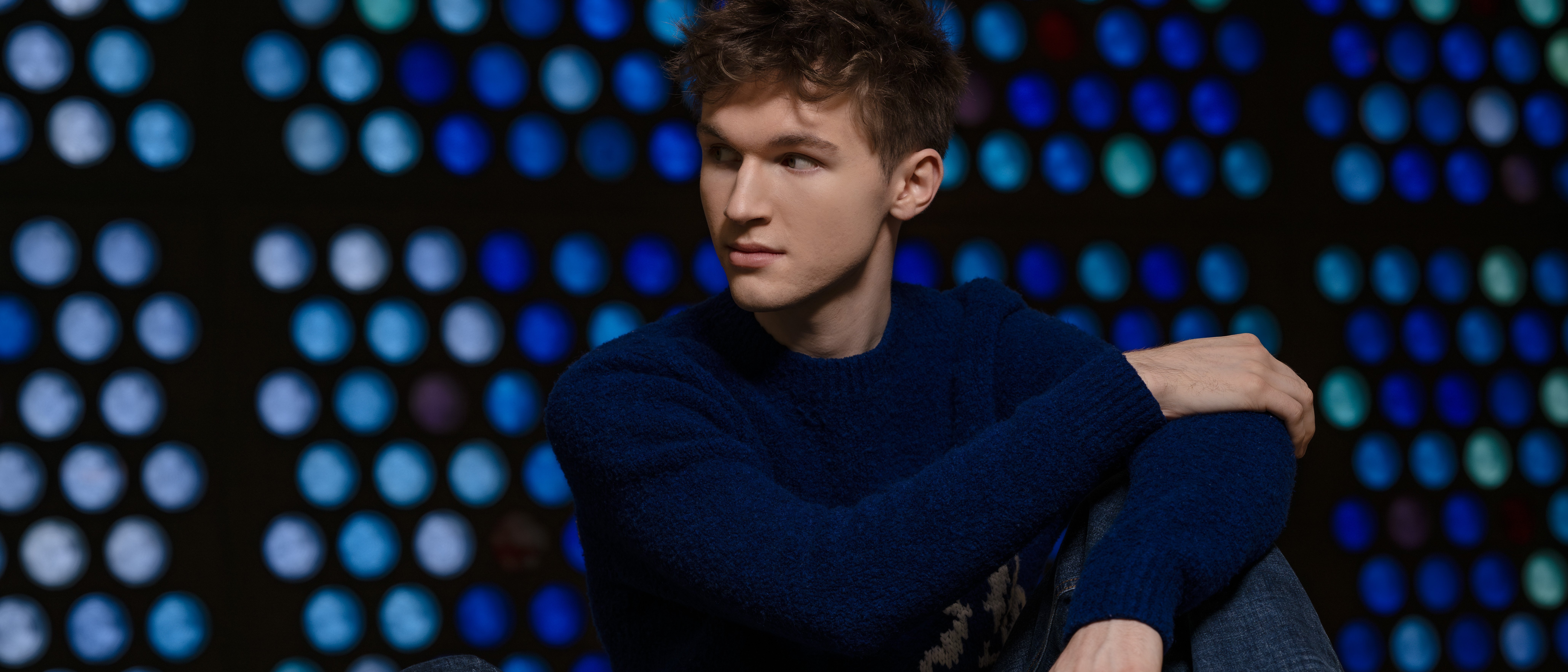 Portrait of the young man Jan Liebermann, sitting in a blue jumper and jeans in front of a background with blue, round glass windows. He is looking to the side. 
