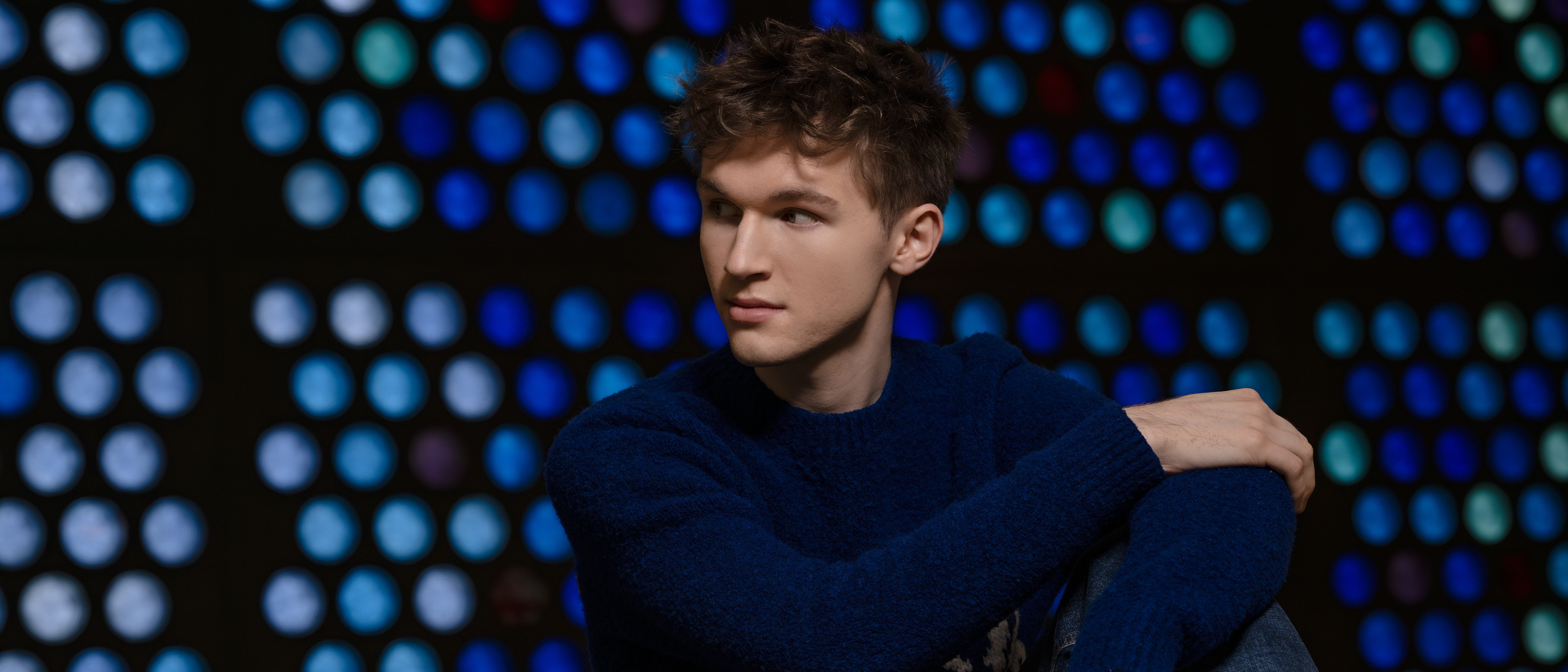 Portrait of the young man Jan Liebermann, sitting in a blue jumper and jeans in front of a background with blue, round glass windows. He is looking to the side. 