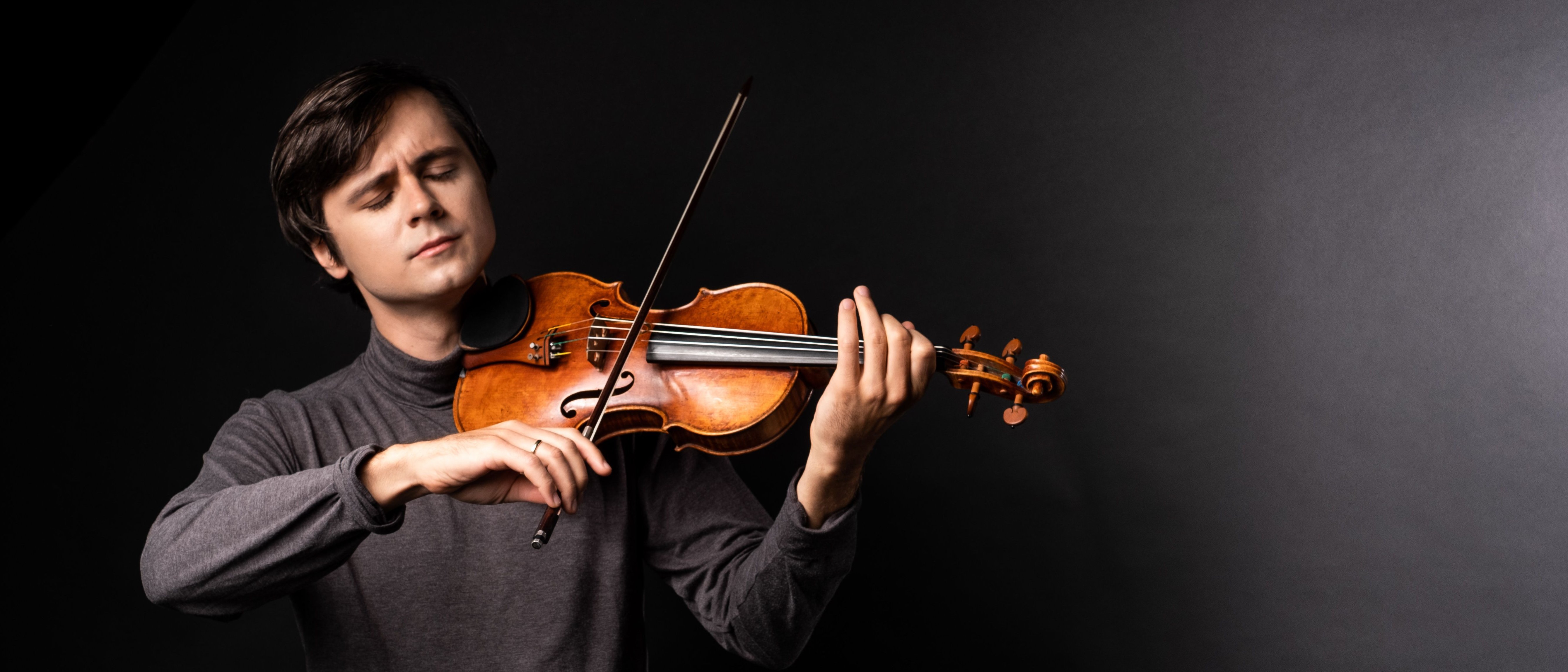 Geiger Aleksey Semenenko mit konzentriertem Blick, dunklem Haar und Violine in der Hand, vor einem stimmungsvollen dunklen Hintergrund.
