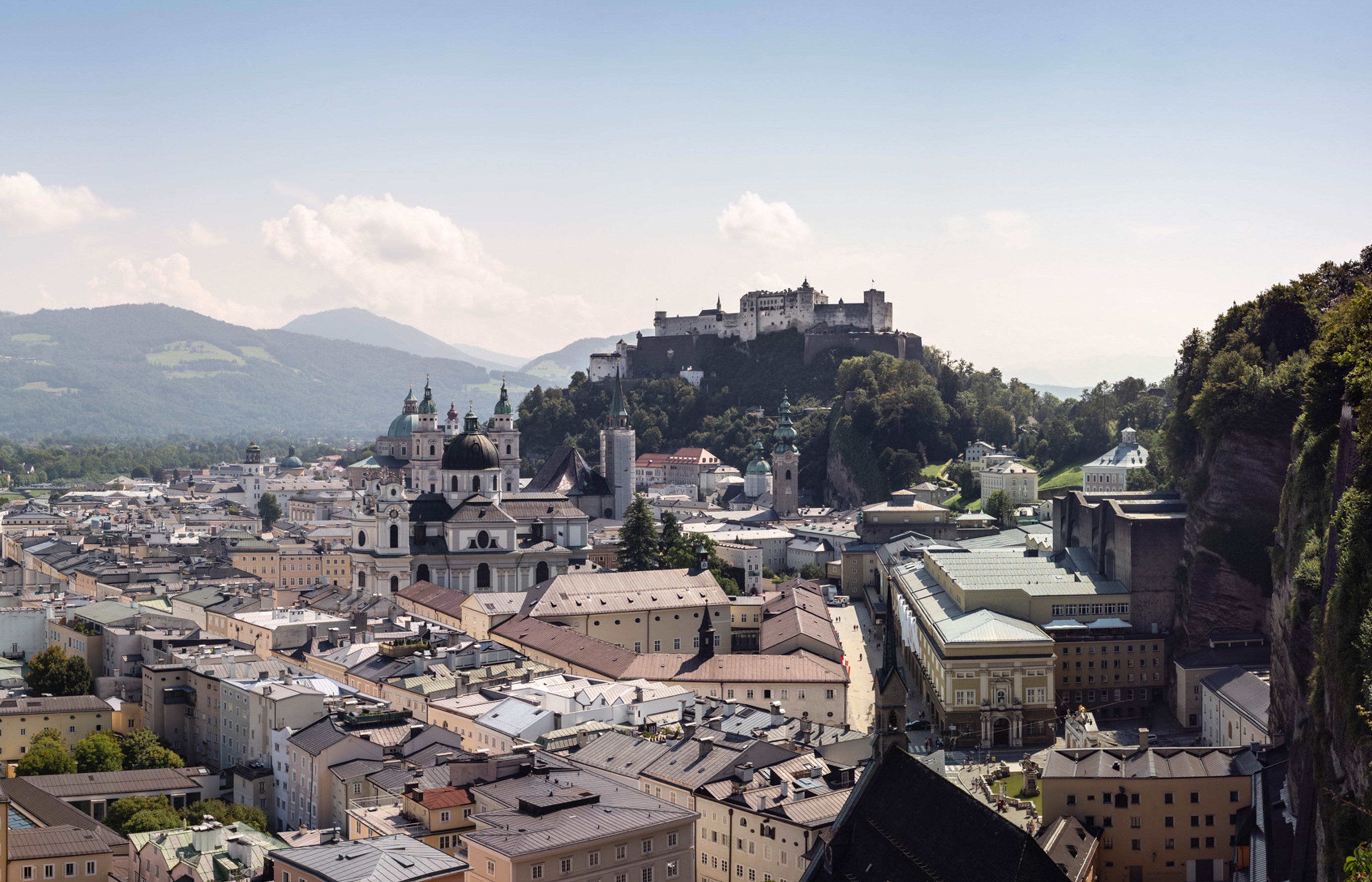 Ein Panoramablick auf Salzburg, Österreich, zeigt eine Mischung aus historischen Gebäuden und moderner Architektur, mit der Festung Hohensalzburg auf einem Hügel. Die Landschaft ist umgeben von üppigen, grünen Hügeln unter klarem Himmel.