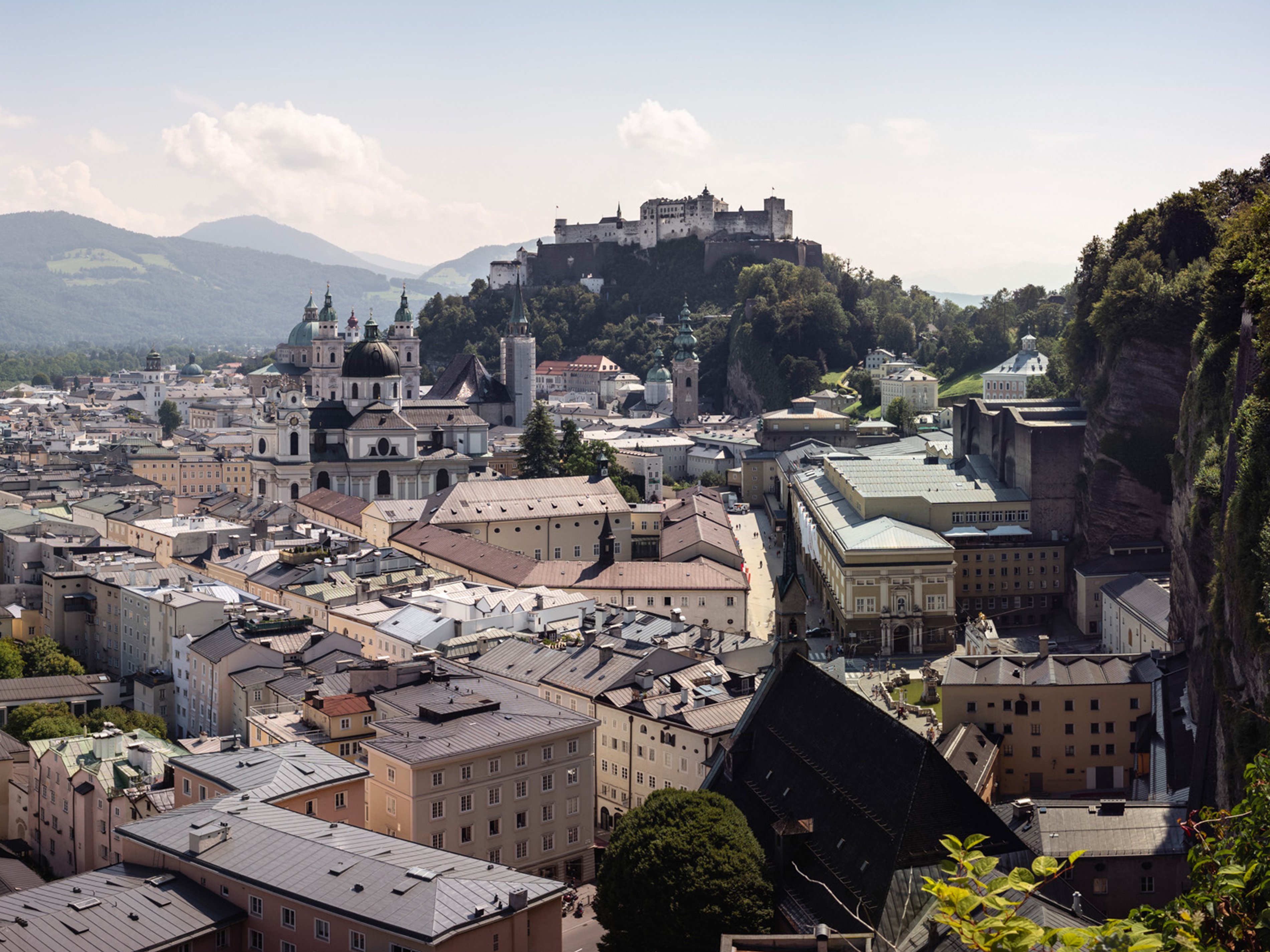 Ein Panoramablick auf Salzburg, Österreich, zeigt eine Mischung aus historischen Gebäuden und moderner Architektur, mit der Festung Hohensalzburg auf einem Hügel. Die Landschaft ist umgeben von üppigen, grünen Hügeln unter klarem Himmel.