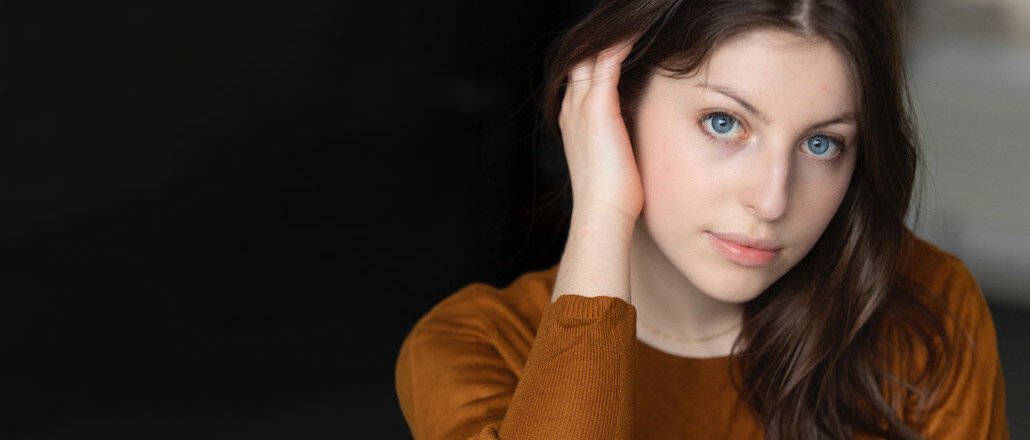 Eine junge Frau mit langen braunen Haaren und blauen Augen, die einen braunen Pullover trägt, sitzt mit einer Hand an ihrem Kopf und blickt sanft in die Kamera vor einem dunklen, verschwommenen Hintergrund.