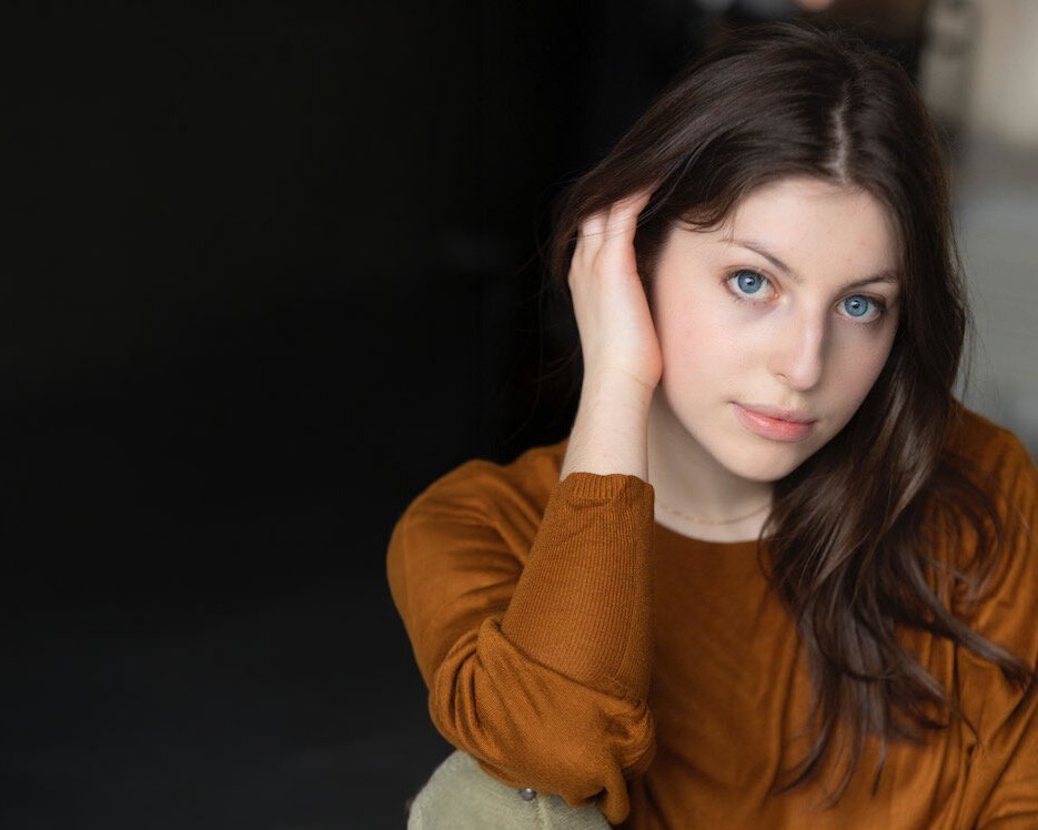 Eine junge Frau mit langen braunen Haaren und blauen Augen, die einen braunen Pullover trägt, sitzt mit einer Hand an ihrem Kopf und blickt sanft in die Kamera vor einem dunklen, verschwommenen Hintergrund.