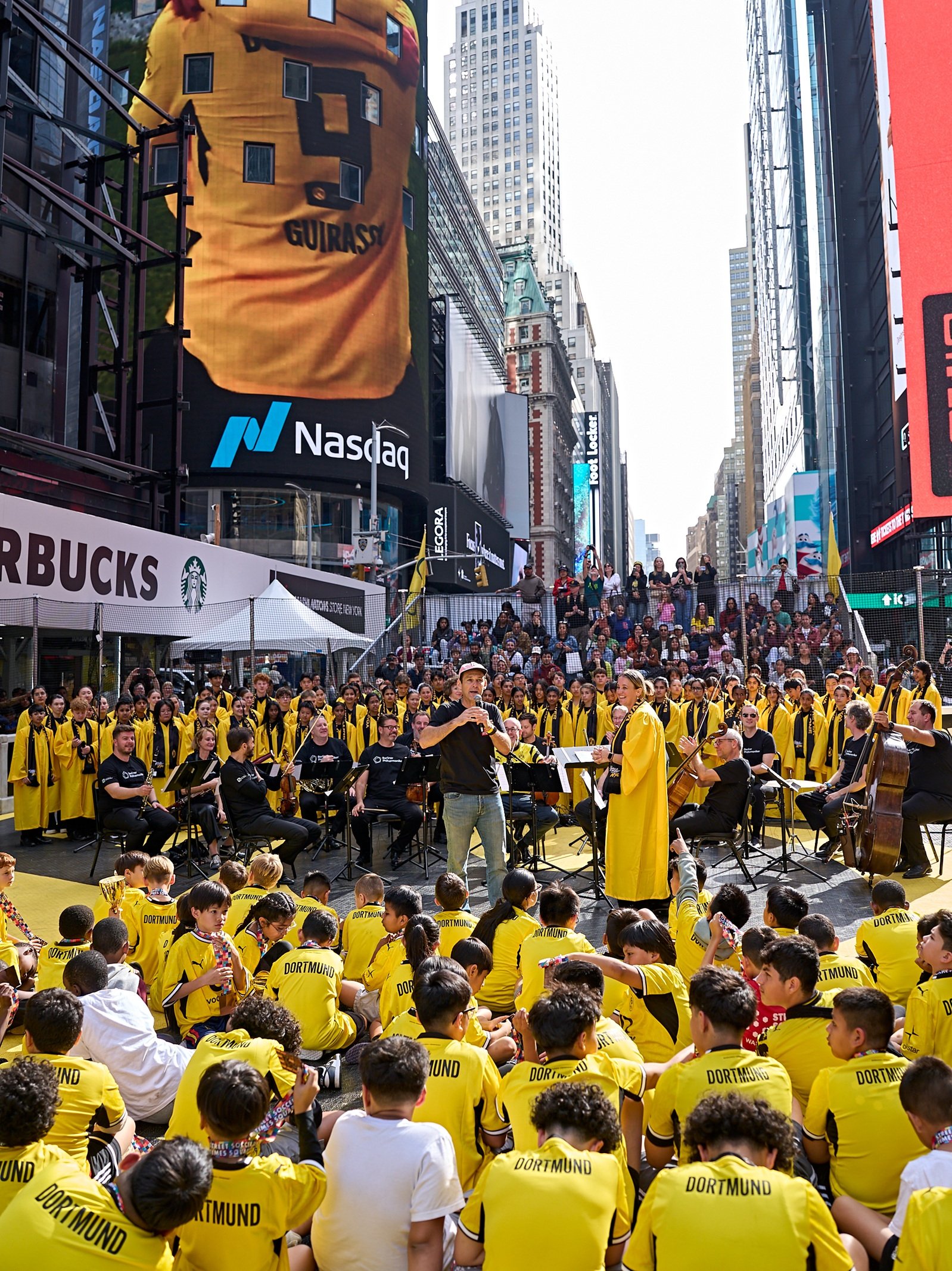 Eine große Gruppe von Kindern in gelben Dortmund-Fußballtrikots sitzt auf dem Boden am Times Square und schaut einem Orchester zu, das unter einer digitalen Werbetafel spielt, auf der das Trikot eines Dortmund-Spielers zu sehen ist.