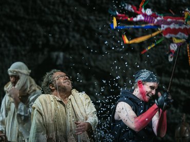 Three people on stage celebrate as one, dressed in black with pink sleeves, breaks a colorful piñata, showering confetti. Another person in beige looks up, smiling, while confetti falls around them.