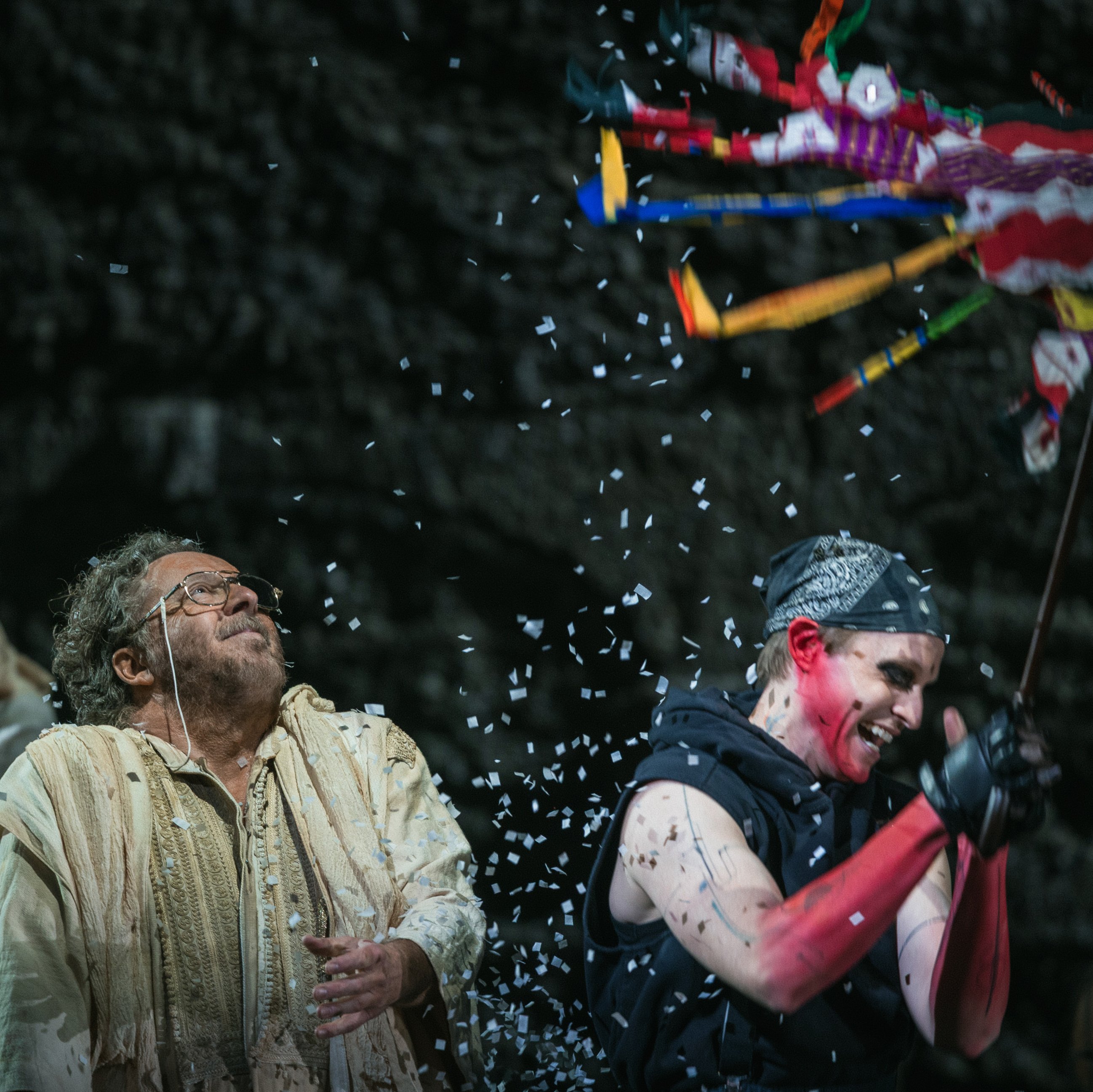 Three people on stage celebrate as one, dressed in black with pink sleeves, breaks a colorful piñata, showering confetti. Another person in beige looks up, smiling, while confetti falls around them.