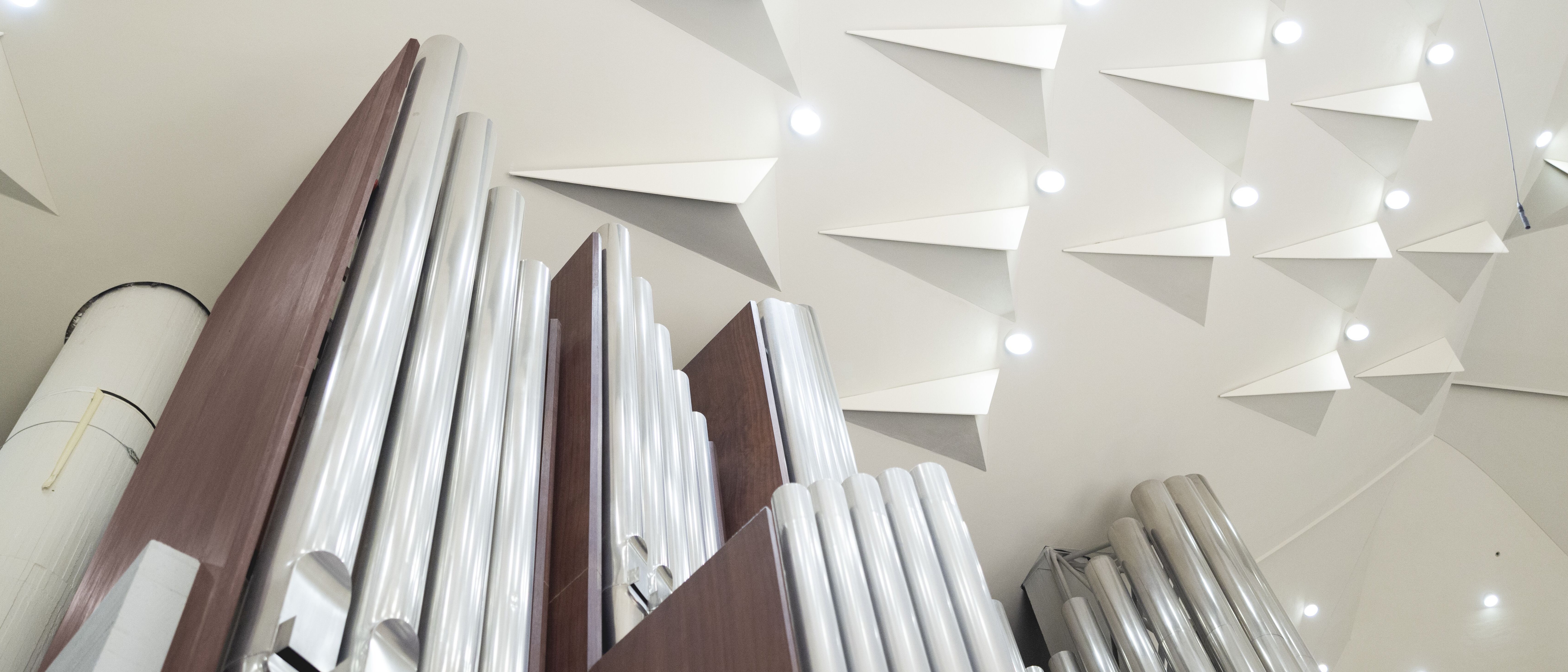 A deep view of gleaming metal organ pipes set in a wooden frame, with the modern ceiling of the Berlin Philharmonie above, featuring geometric white panels and circular lights.