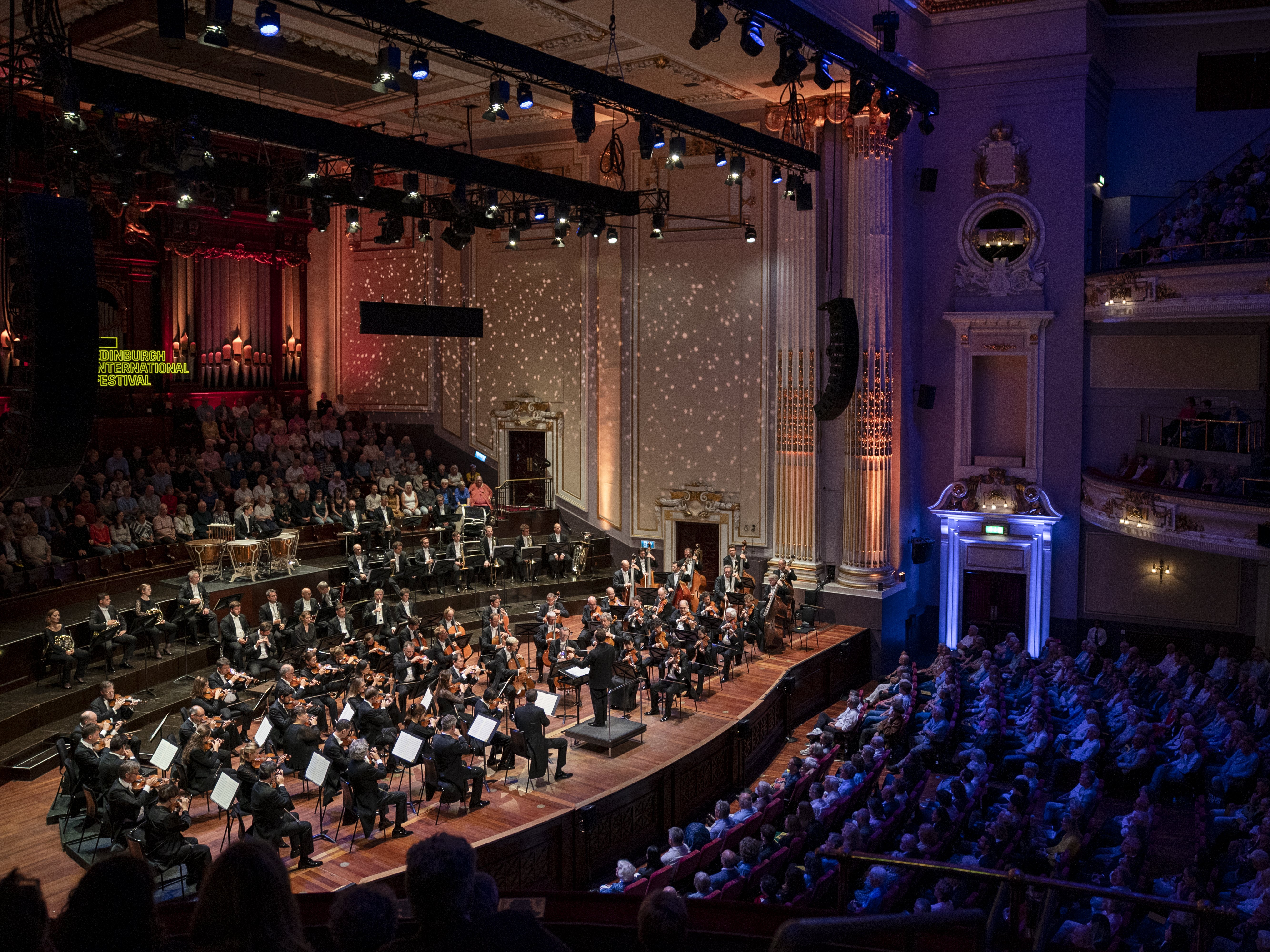 A full orchestra performs onstage in a grand concert hall filled with an audience. Warm lighting highlights the musicians, while soft spotlights create patterns on the walls.