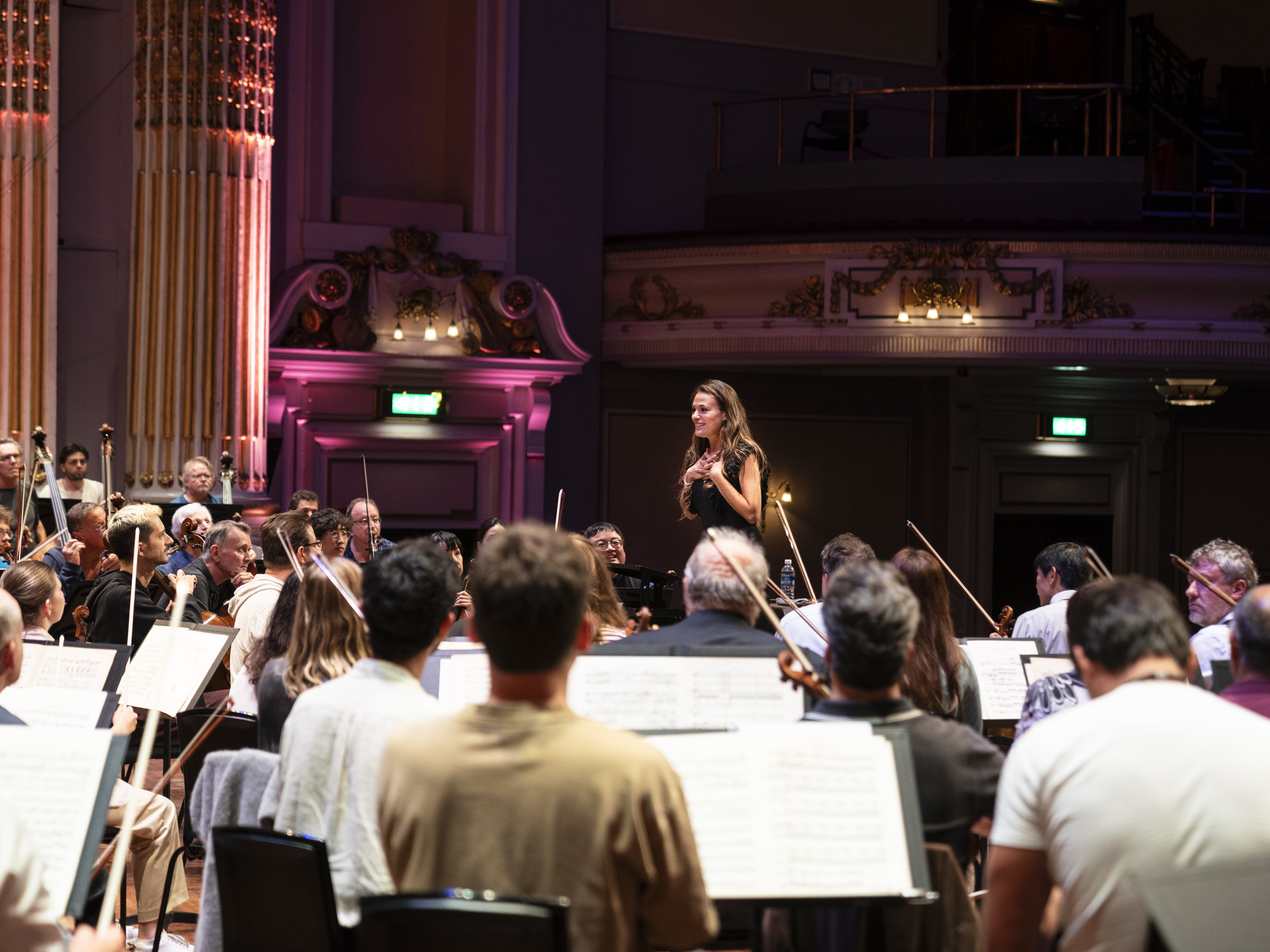 A woman stands in front of an orchestra during rehearsal in a grand concert hall with ornate decor and sheet music visible on music stands.