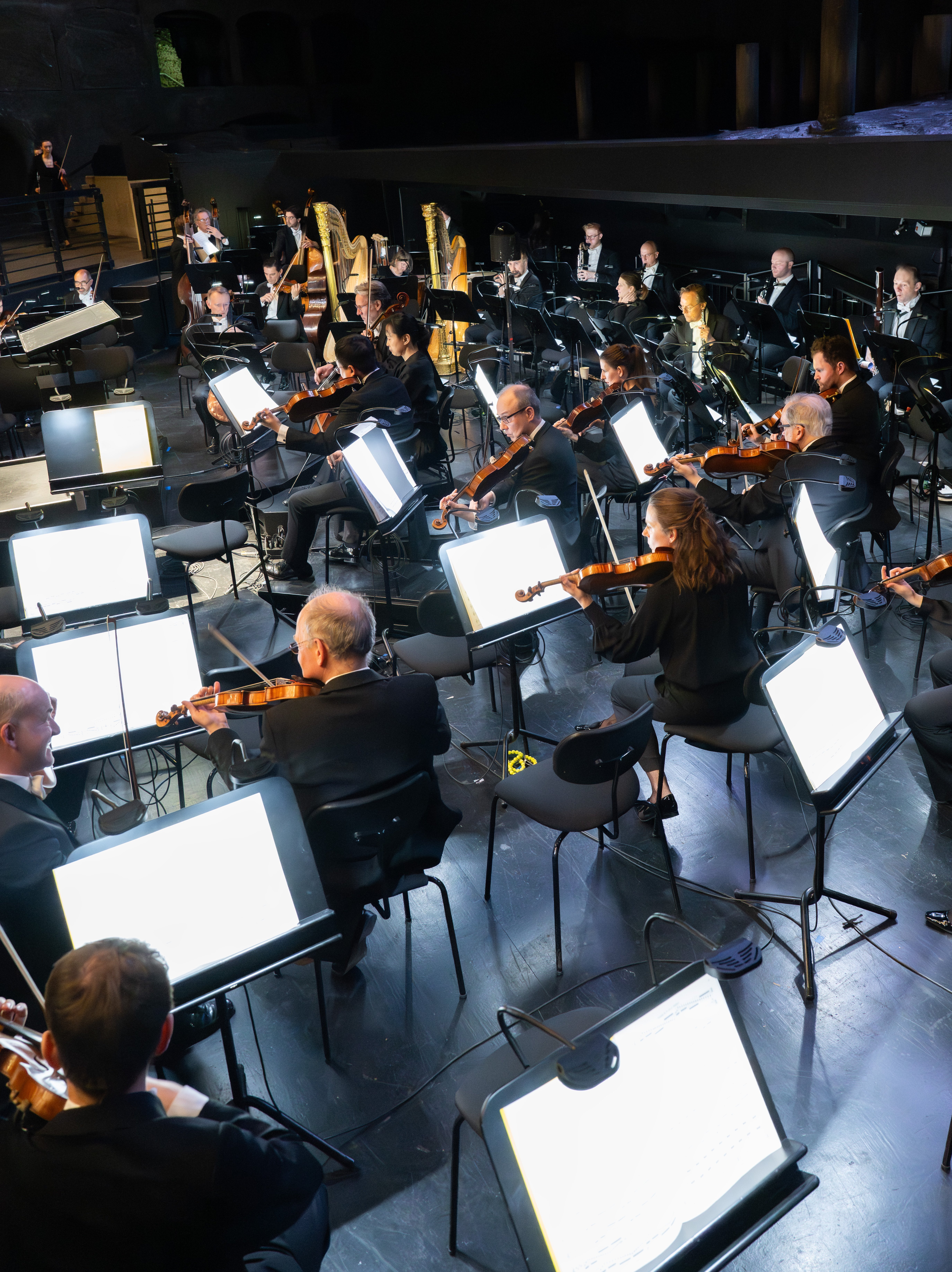 Musiker*innen im Orchestergraben, die Streich-, Blech- und Blasinstrumente spielen. Ihre Notenständer sind beleuchtet und bilden einen hellen Kontrast zum dunklen Hintergrund.