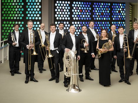 Eine Gruppe von 12 Musikern in formeller schwarzer Kleidung steht in einer Halle und hält Blechblasinstrumente, darunter Posaunen, Trompeten, eine Tuba und Waldhörner, vor einer bunt gepunkteten Wand im Hintergrund.