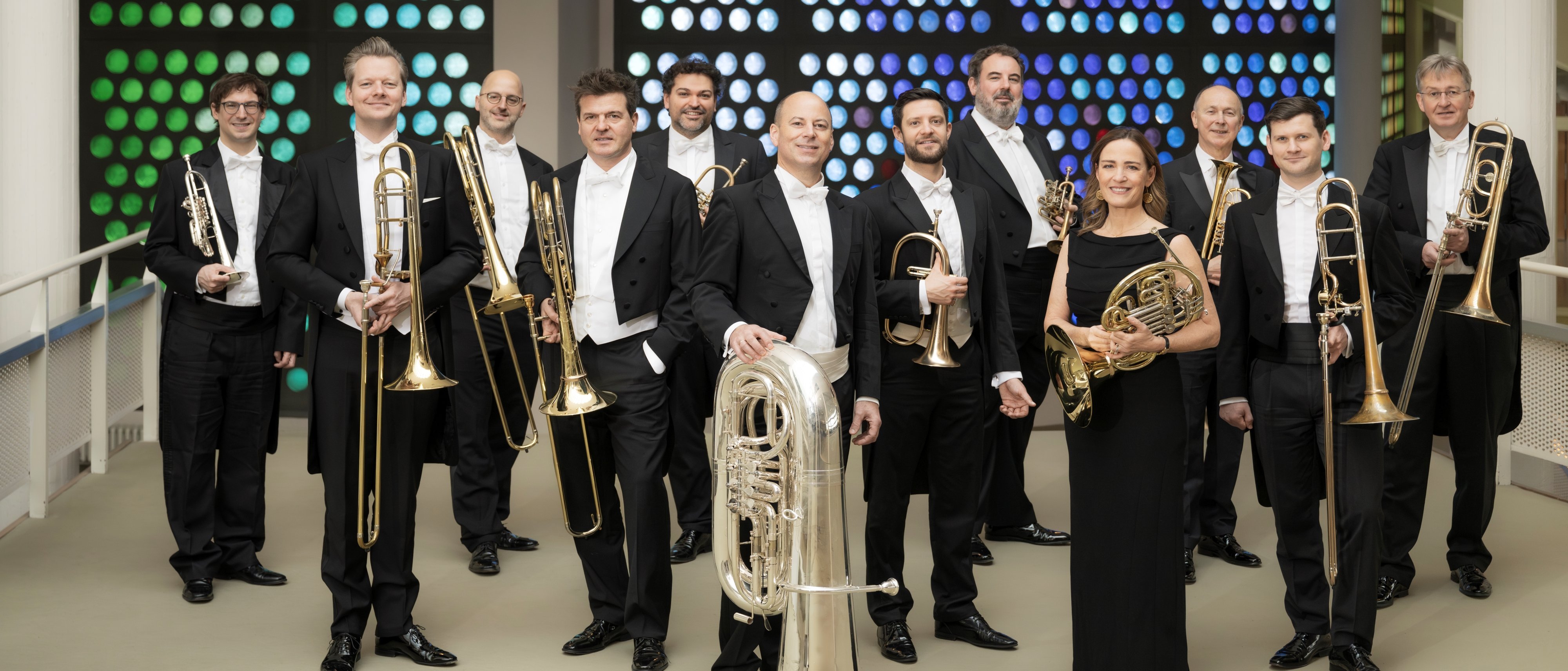 Eine Gruppe von 12 Musikern in formeller schwarzer Kleidung steht in einer Halle und hält Blechblasinstrumente, darunter Posaunen, Trompeten, eine Tuba und Waldhörner, vor einer bunt gepunkteten Wand im Hintergrund.