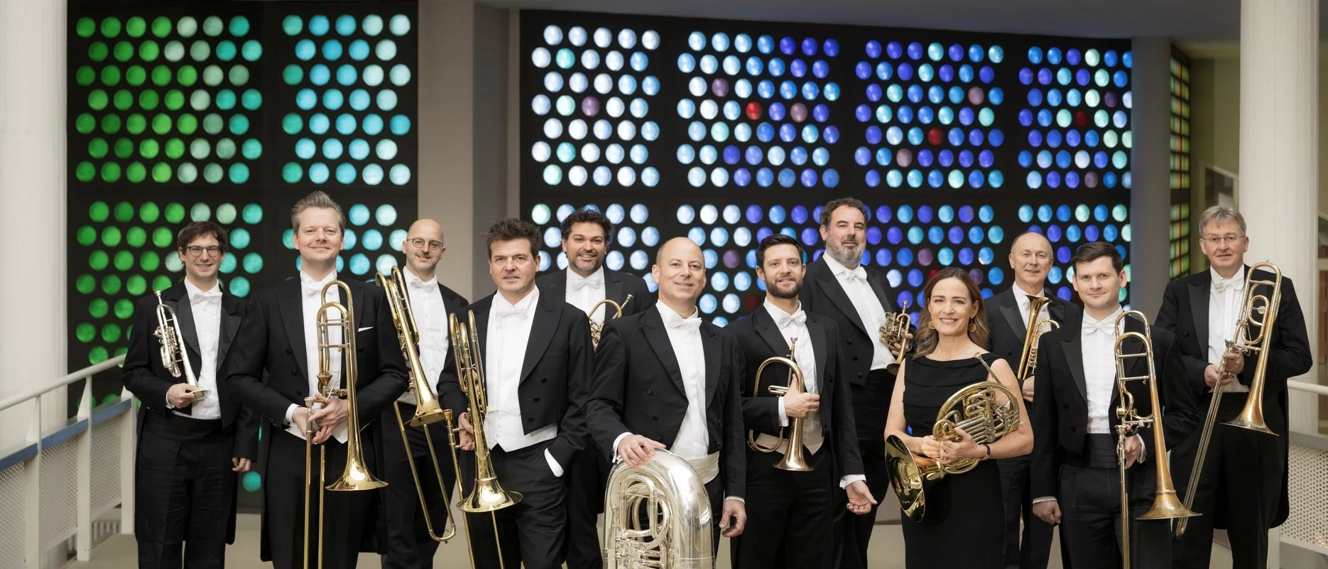 Eine Gruppe von 12 Musikern in formeller schwarzer Kleidung steht in einer Halle und hält Blechblasinstrumente, darunter Posaunen, Trompeten, eine Tuba und Waldhörner, vor einer bunt gepunkteten Wand im Hintergrund.