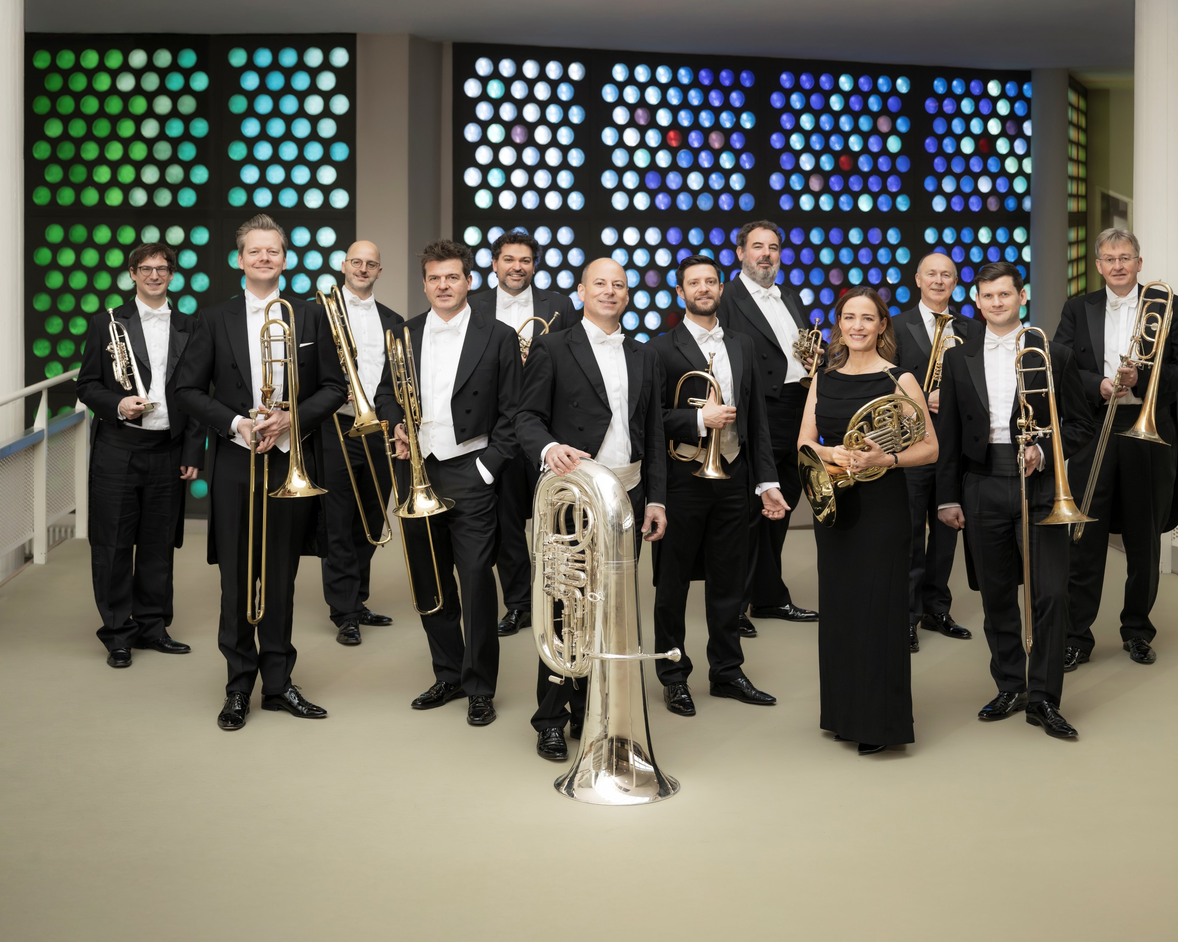 Eine Gruppe von 12 Musikern in formeller schwarzer Kleidung steht in einer Halle und hält Blechblasinstrumente, darunter Posaunen, Trompeten, eine Tuba und Waldhörner, vor einer bunt gepunkteten Wand im Hintergrund.