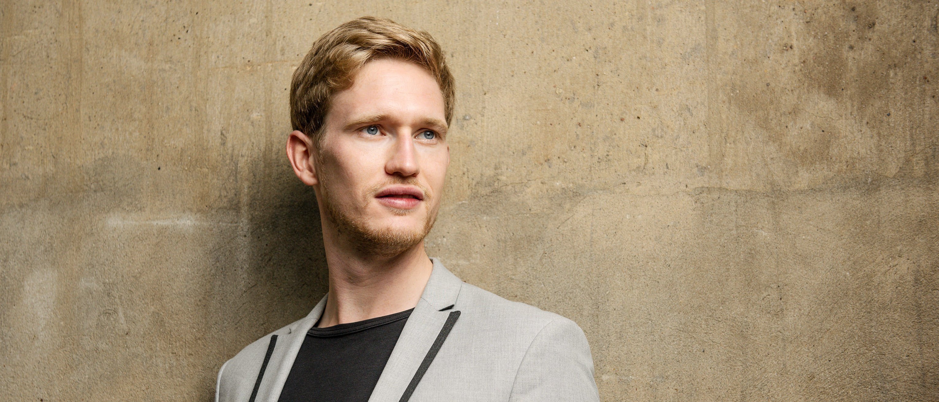 Young man with blond hair and a light beard, wearing a light grey jacket with a black shirt, in front of a textured concrete wall. He looks slightly to the side.