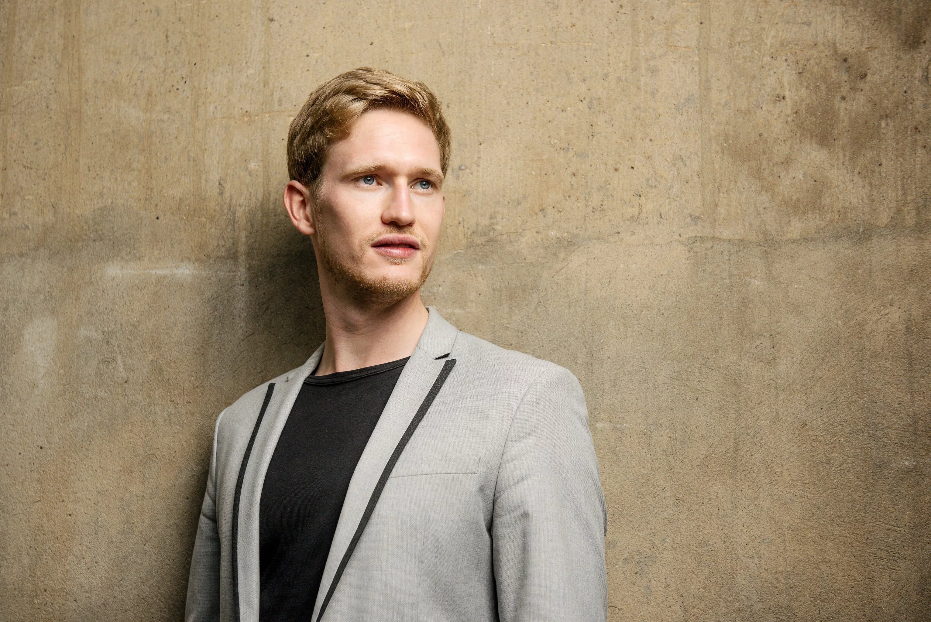 Junger Mann mit blondem Haar und leichtem Bart, in einem hellgrauen Sakko mit schwarzem Shirt, vor einer strukturierten Betonwand. Sein Blick geht leicht zur Seite.
