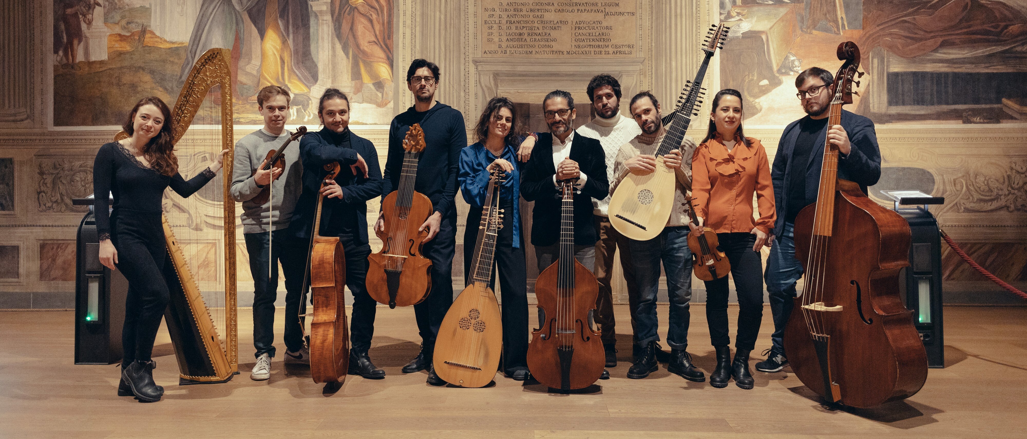Elf Musiker posieren in einem Innenraum mit klassischen Streich- und Zupfinstrumenten, darunter eine Harfe, Geigen und Lauten, vor einer dekorativen Wand mit Wandmalereien und Inschriften.