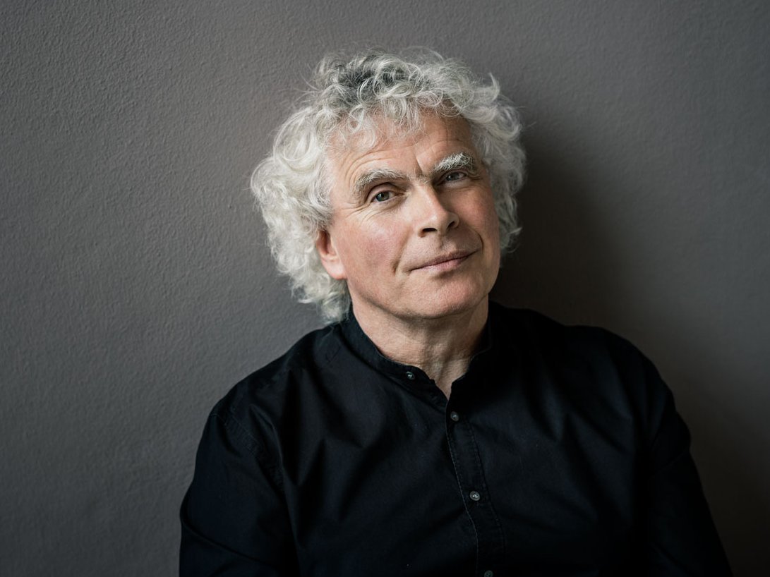 Ein Mann mit weißen Locken und einem schwarzen Hemd steht vor einer neutralen Wand. Sein Blick in die Kamera ist entspannt.