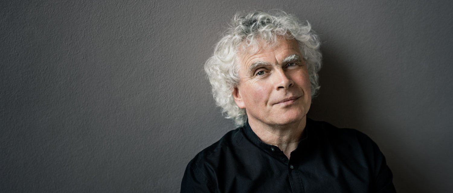 Ein Mann mit weißen Locken und einem schwarzen Hemd steht vor einer neutralen Wand. Sein Blick in die Kamera ist entspannt.