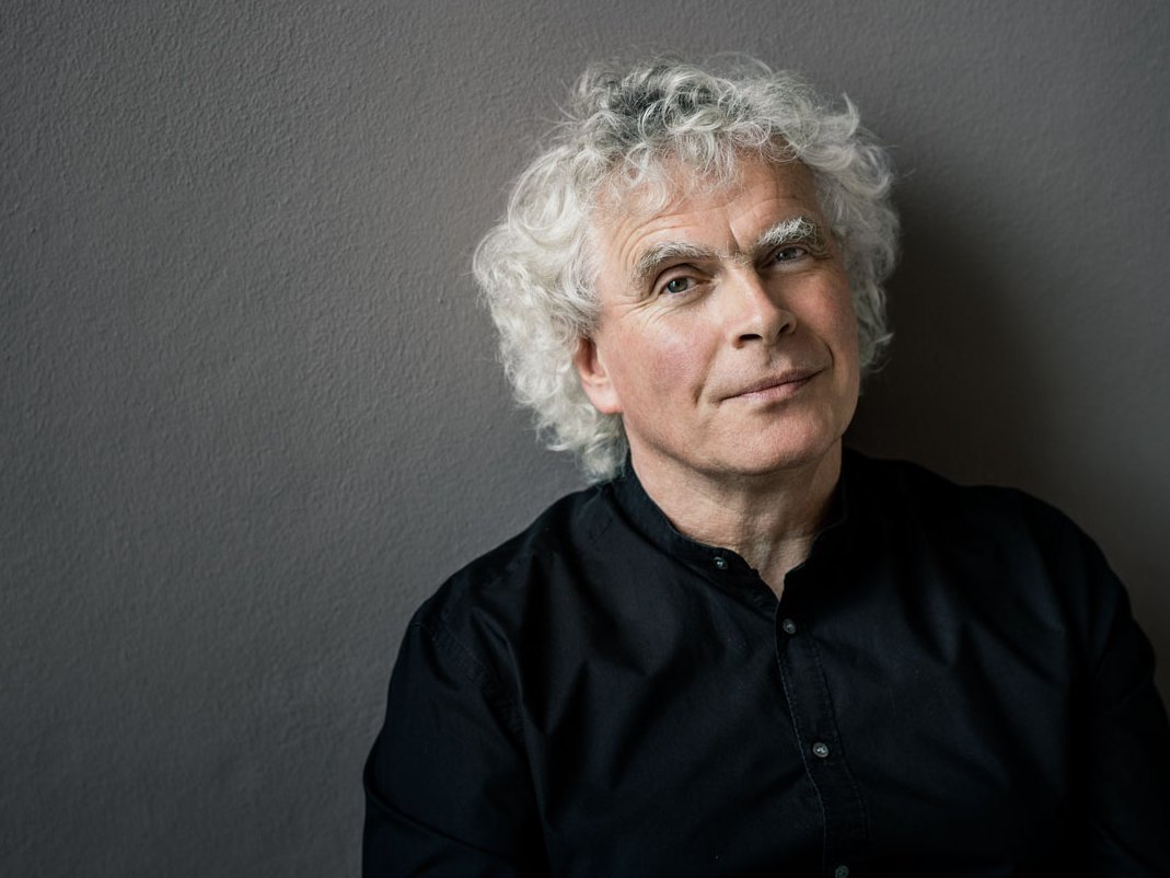 Ein Mann mit weißen Locken und einem schwarzen Hemd steht vor einer neutralen Wand. Sein Blick in die Kamera ist entspannt.
