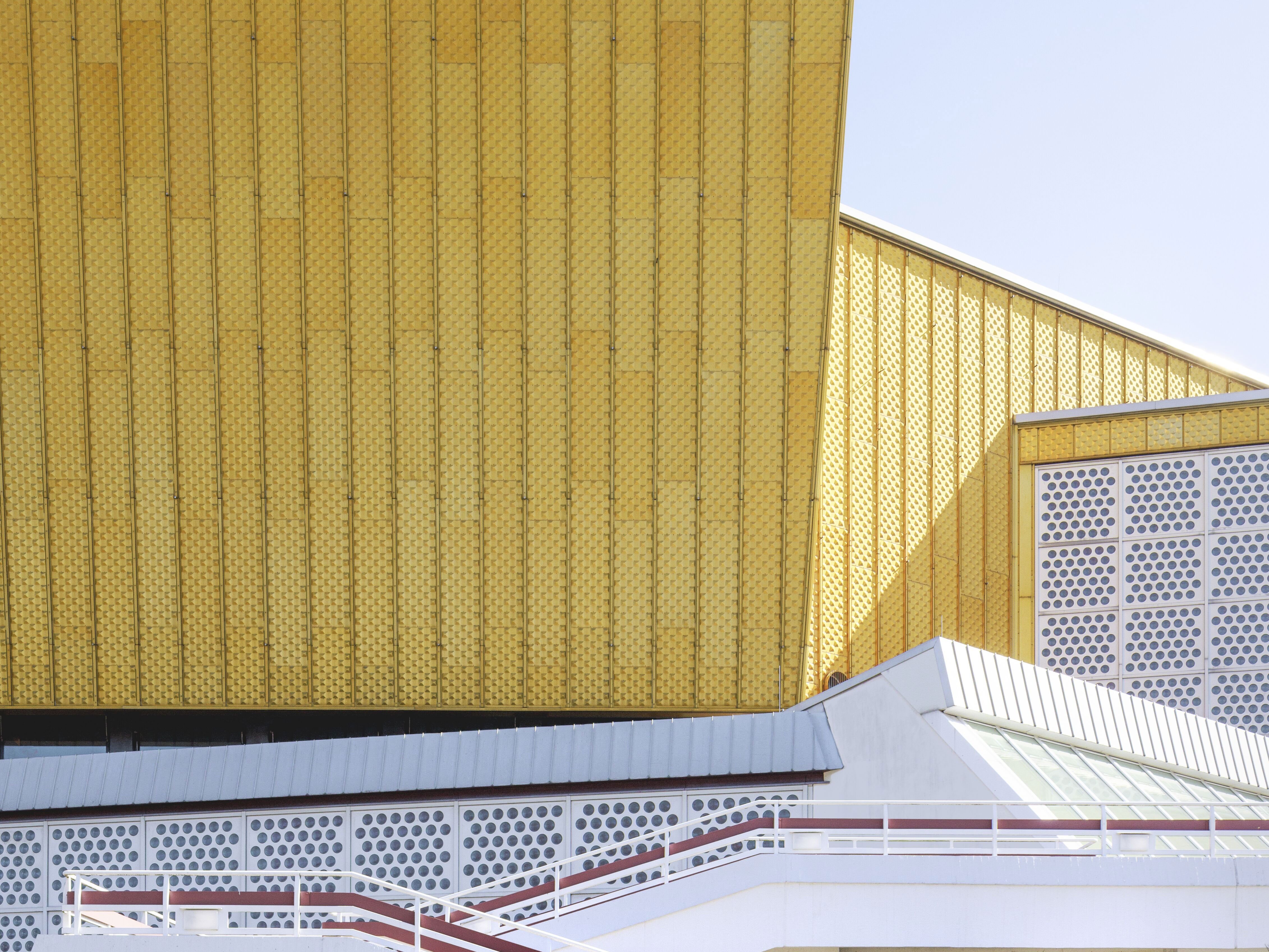 Gelbes, modernes Gebäude mit eckigem Design und weißen, abgestuften Rampen im Vordergrund. Die Struktur bildet einen Kontrast zum klaren blauen Himmel und schafft eine beeindruckende architektonische Szene.