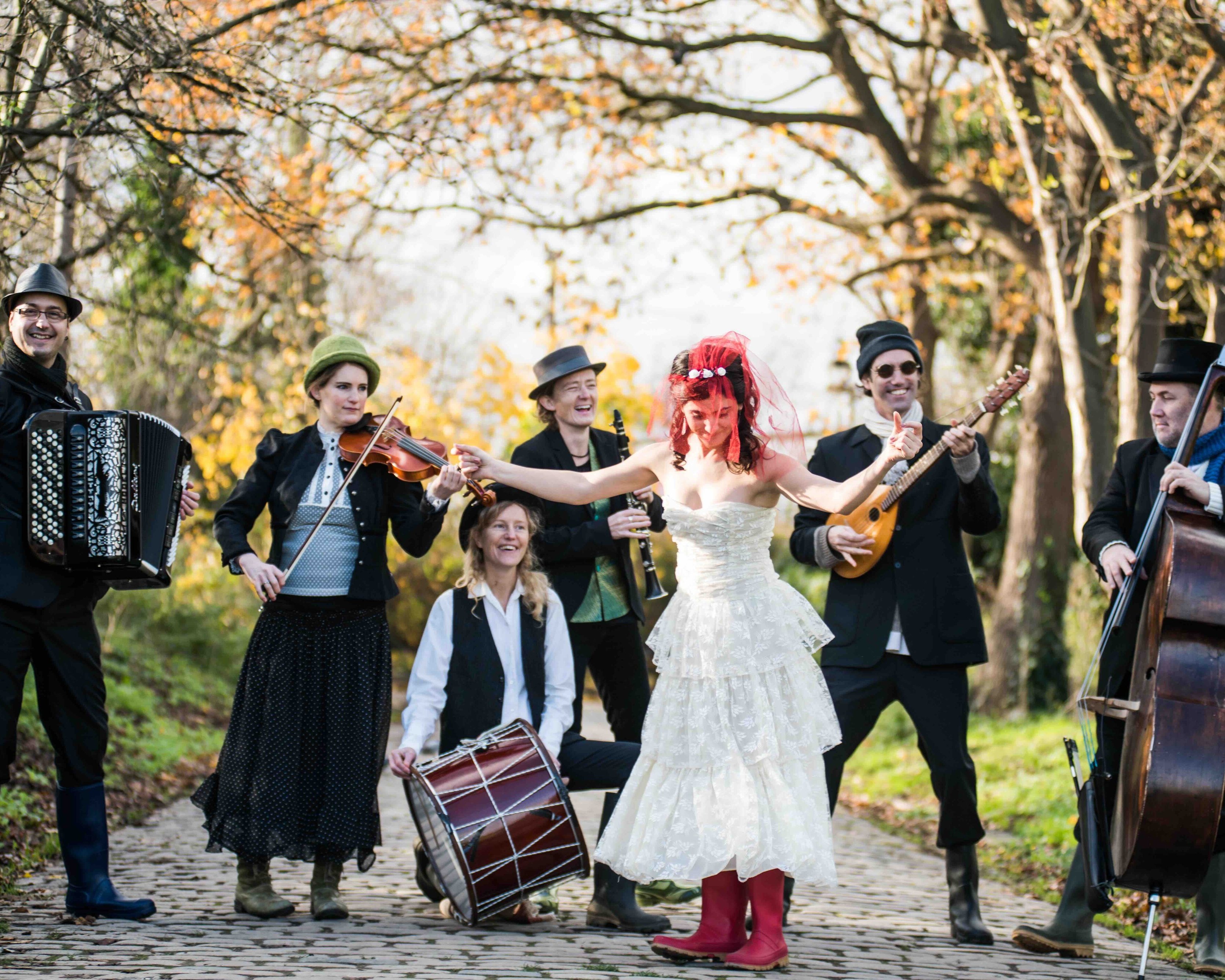 Eine Gruppe von sechs Musikern spielt im Freien auf einem Kopfsteinpflasterweg, im Hintergrund sind herbstlich gefärbte Bäume zu sehen. Eine Person trägt ein weißes Kleid und rote Stiefel, hält eine Gitarre in der Hand und singt.