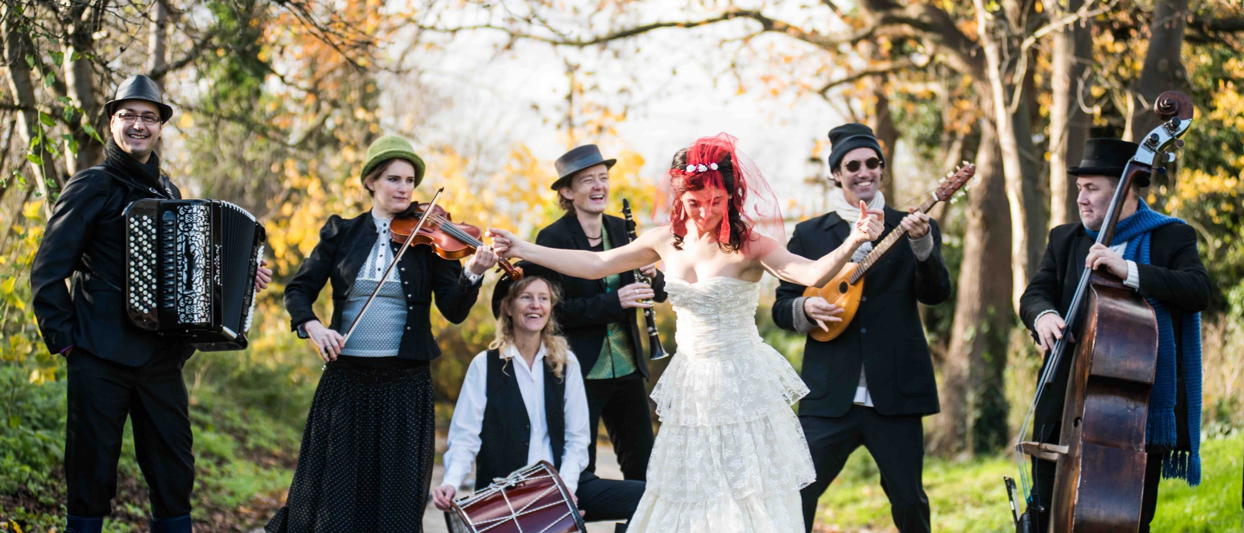 Eine Gruppe von sechs Musikern spielt im Freien auf einem Kopfsteinpflasterweg, im Hintergrund sind herbstlich gefärbte Bäume zu sehen. Eine Person trägt ein weißes Kleid und rote Stiefel, hält eine Gitarre in der Hand und singt.