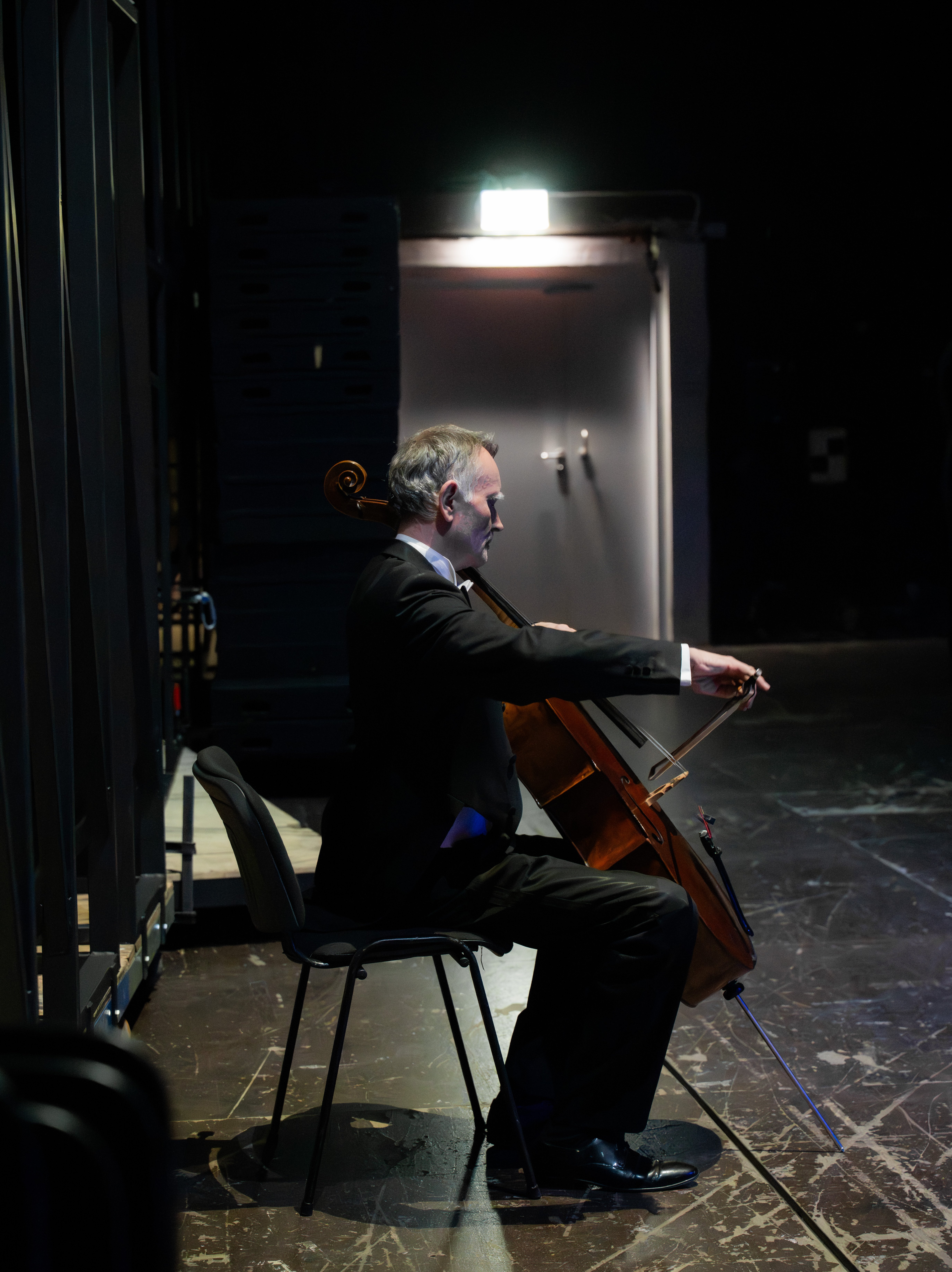 Ein Mann im Anzug sitzt auf einem Stuhl hinter der Bühne und spielt ein Cello in einem schwach beleuchteten Raum mit einem hellen Ausgangsschild und einer Tür im Hintergrund.