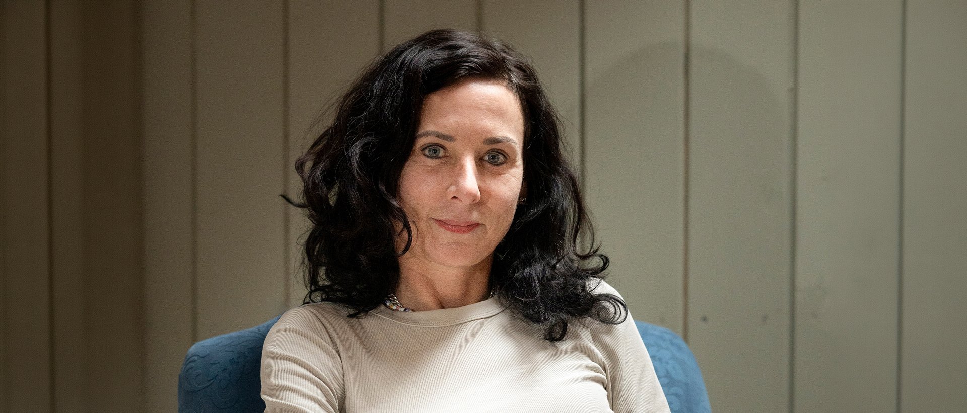 Eine Frau mit gewelltem dunklem Haar und einem hellbeigen, langärmeligen Hemd sitzt auf einem blau gemusterten Stuhl und blickt in die Kamera. Sie hat einen neutralen Gesichtsausdruck und eine Holzverkleidung im Hintergrund.