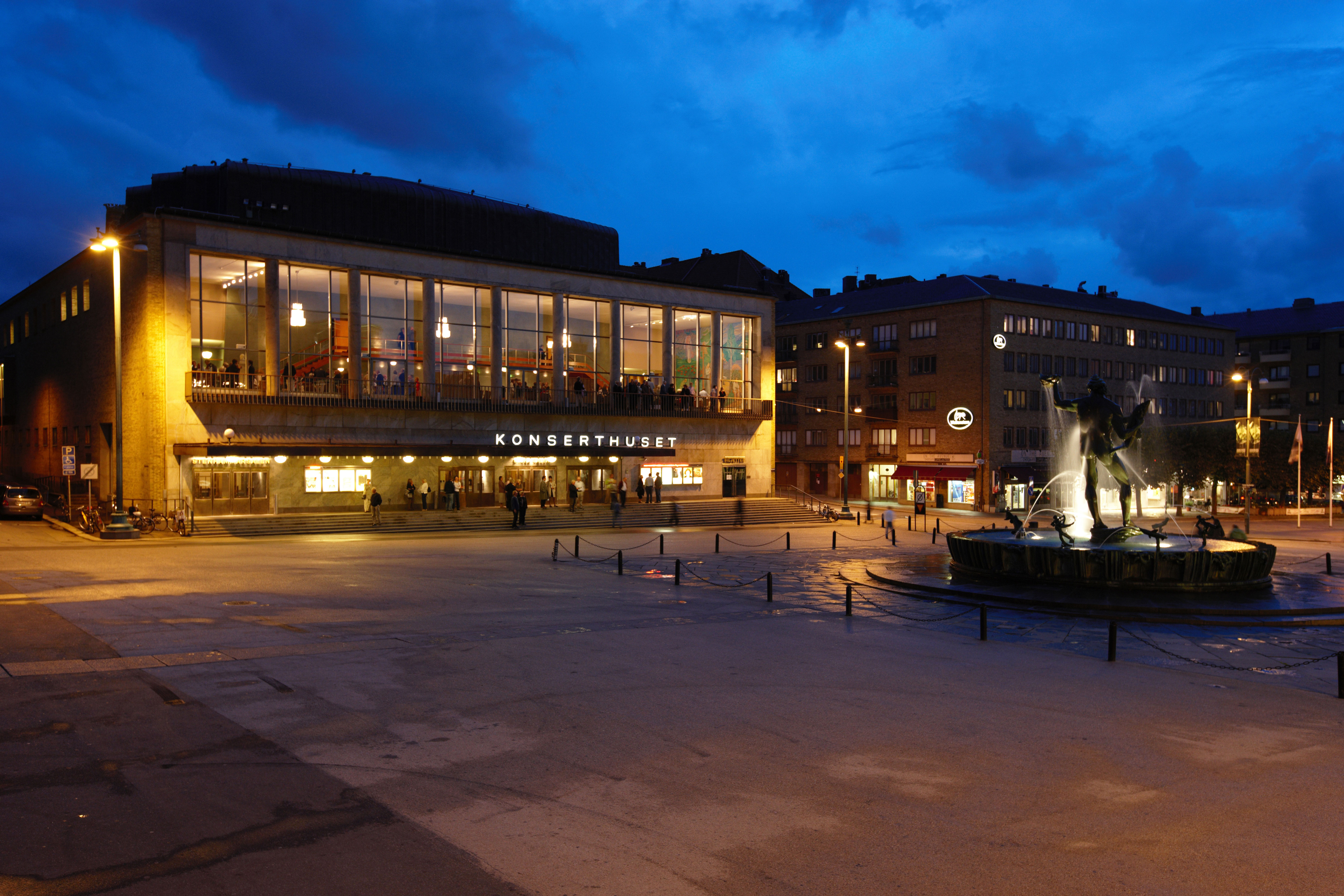 A concert hall with large windows, warmly lit from inside, stands next to a square featuring a fountain with a statue, all under a deep blue evening sky.