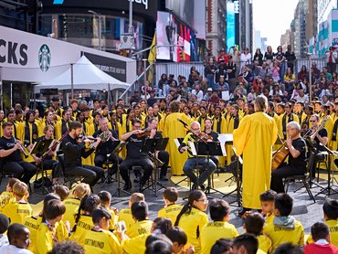 Ein Orchester, dessen Mitglieder größtenteils gelbe T-Shirts tragen, spielt auf dem Times Square vor einem großen Publikum aus Kindern, die ebenfalls gelbe T-Shirts tragen.