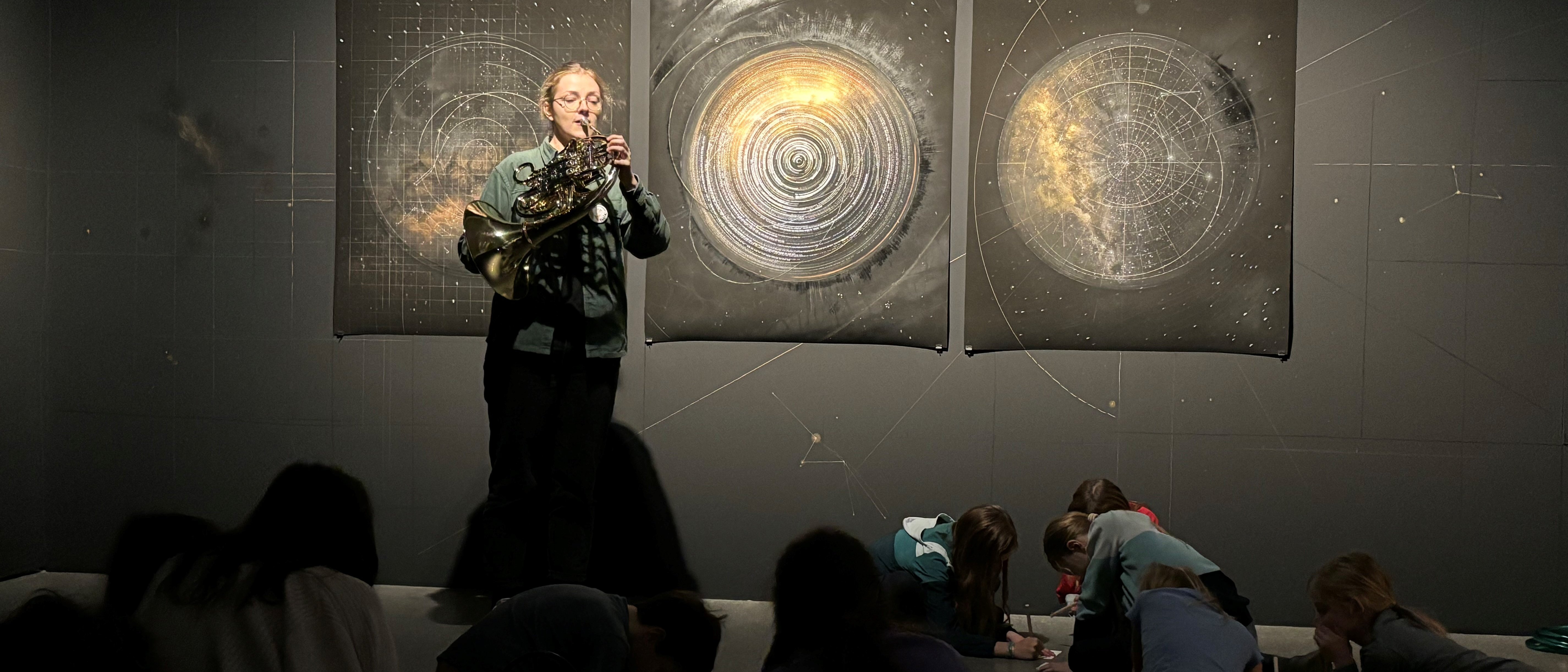 Hornistin Paula Ernesaks spielt vor drei kreisförmigen Kunstwerken an der Wand, die den Weltraum thematisieren, während eine Gruppe von Kindern auf dem Boden sitzt und bei gedämpftem Licht zeichnet oder schreibt.