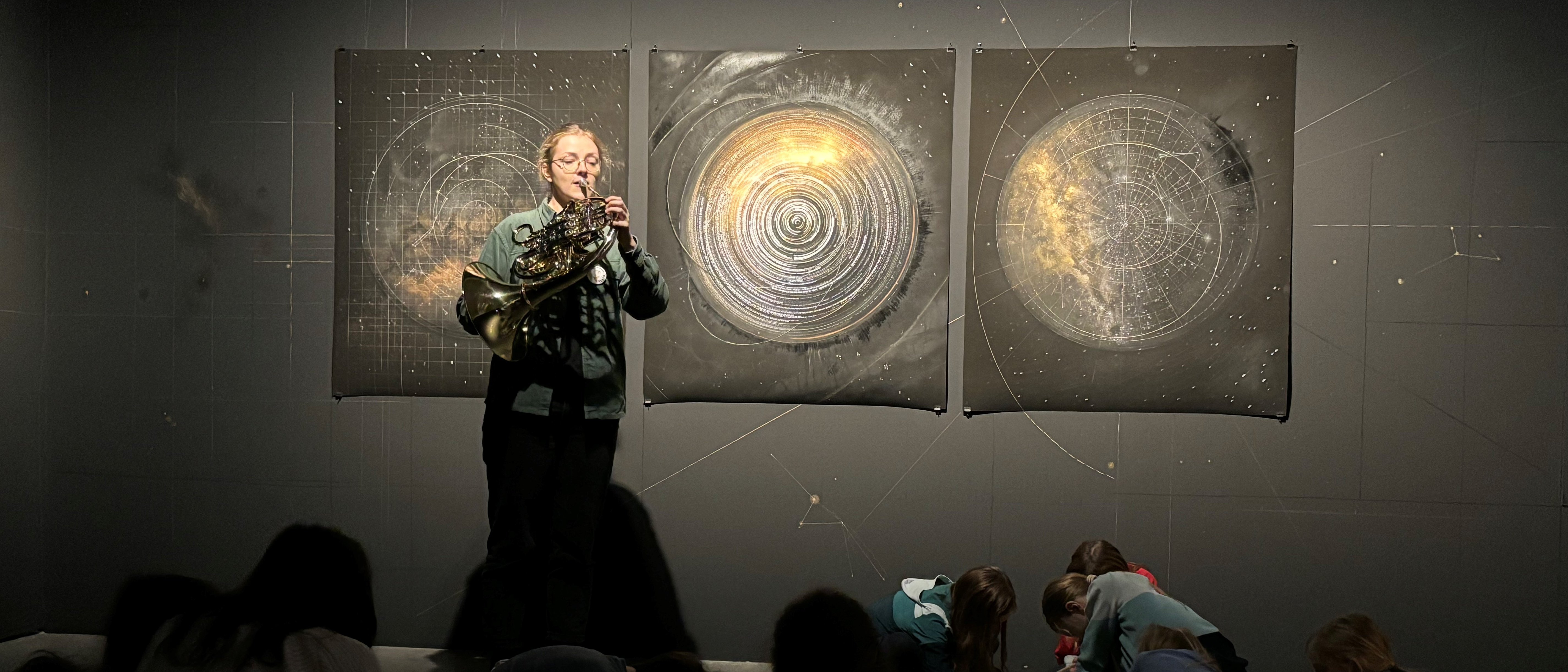 Horn player Paula Ernesaks plays in front of three circular artworks on the wall depicting outer space, while a group of children sit on the floor drawing or writing in subdued lighting.