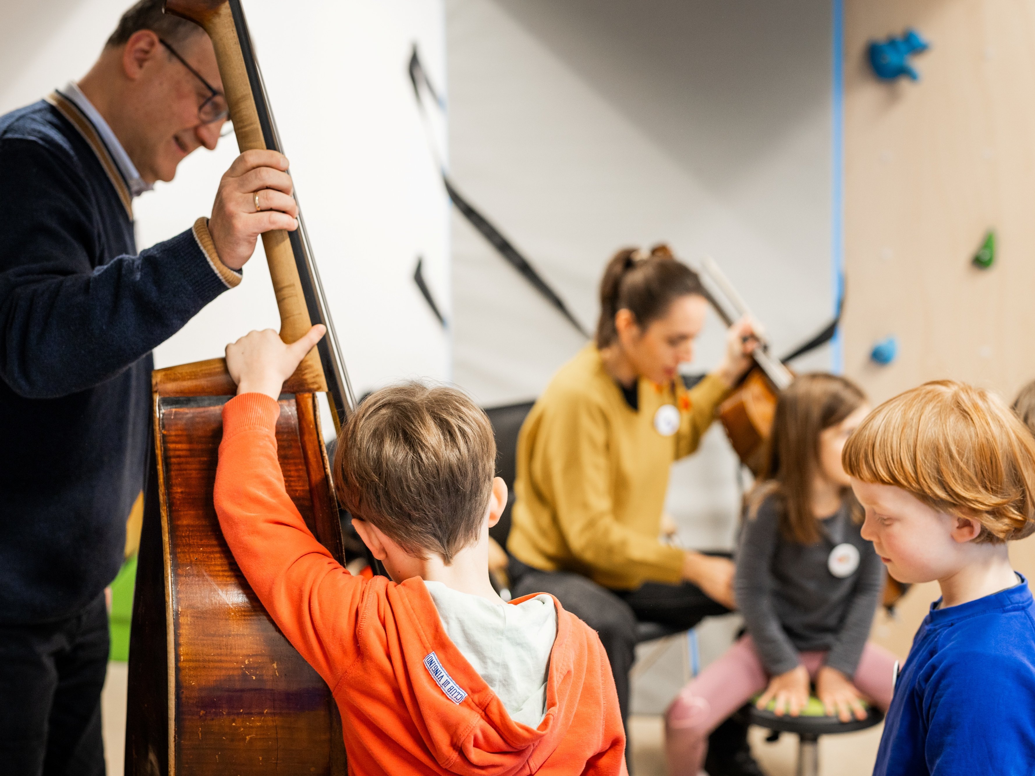 Ein Mann spielt einen Kontrabass, während eine Gruppe von Kindern aufmerksam zuhört. Im Hintergrund spielt eine Frau auf einem Cello. Die Kinder wirken engagiert und interessiert an der Musik.