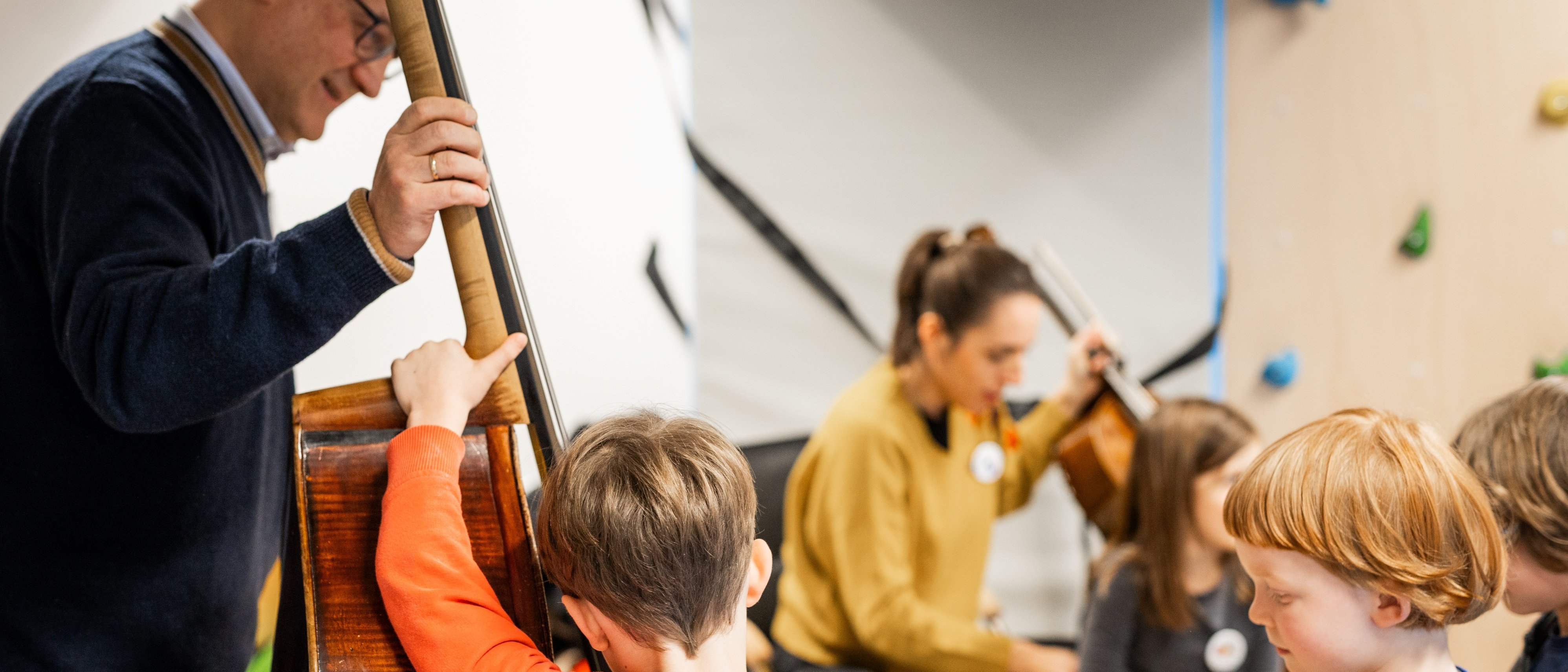 Ein Mann spielt einen Kontrabass, während eine Gruppe von Kindern aufmerksam zuhört. Im Hintergrund spielt eine Frau auf einem Cello. Die Kinder wirken engagiert und interessiert an der Musik.