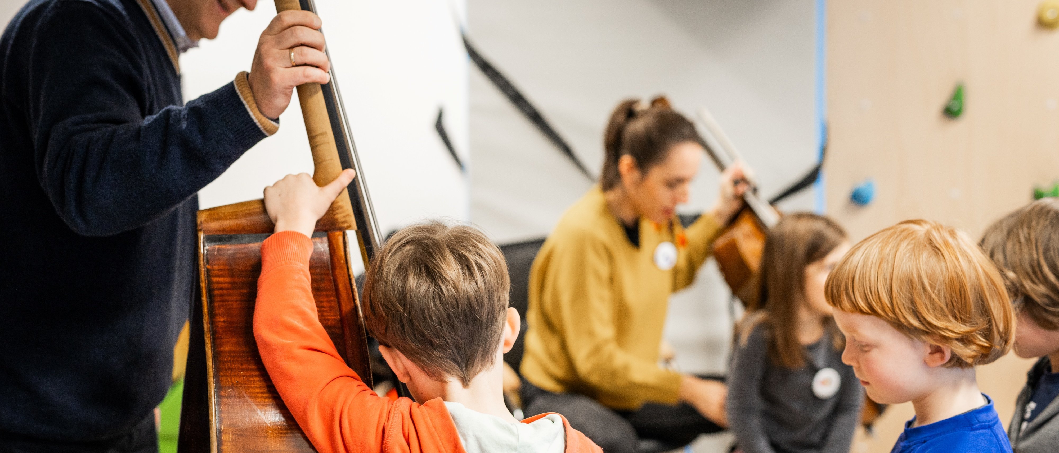 Ein Mann spielt einen Kontrabass, während eine Gruppe von Kindern aufmerksam zuhört. Im Hintergrund spielt eine Frau auf einem Cello. Die Kinder wirken engagiert und interessiert an der Musik.