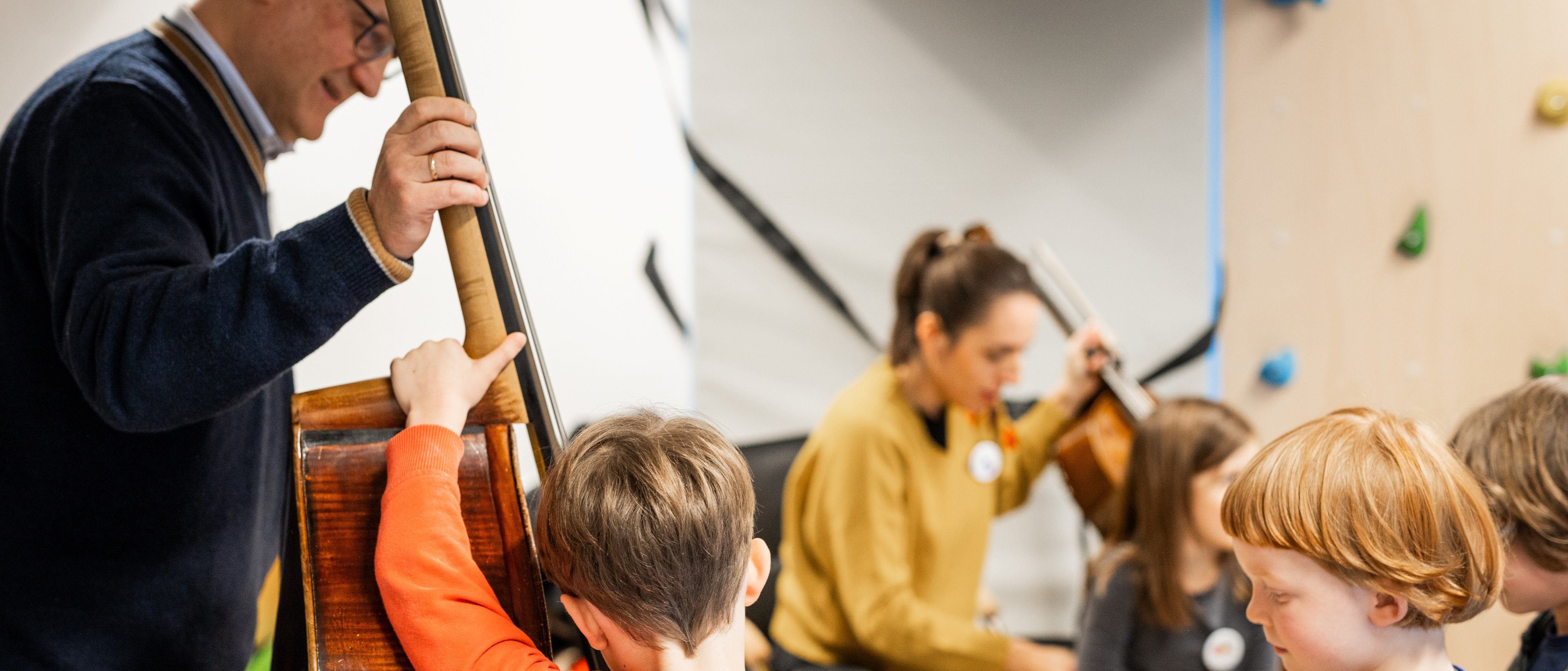 Ein Mann spielt einen Kontrabass, während eine Gruppe von Kindern aufmerksam zuhört. Im Hintergrund spielt eine Frau auf einem Cello. Die Kinder wirken engagiert und interessiert an der Musik.