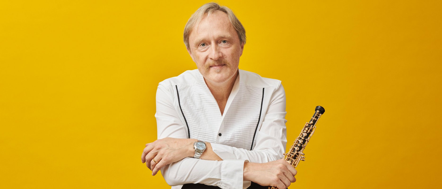 Ein Mann mit hellem Haar und Schnurrbart, bekleidet mit einem weißen Hemd, sitzt vor einem leuchtend gelben Hintergrund, hält eine Oboe in der Hand und stützt seine Arme auf die Lehne eines schwarzen Stuhls.