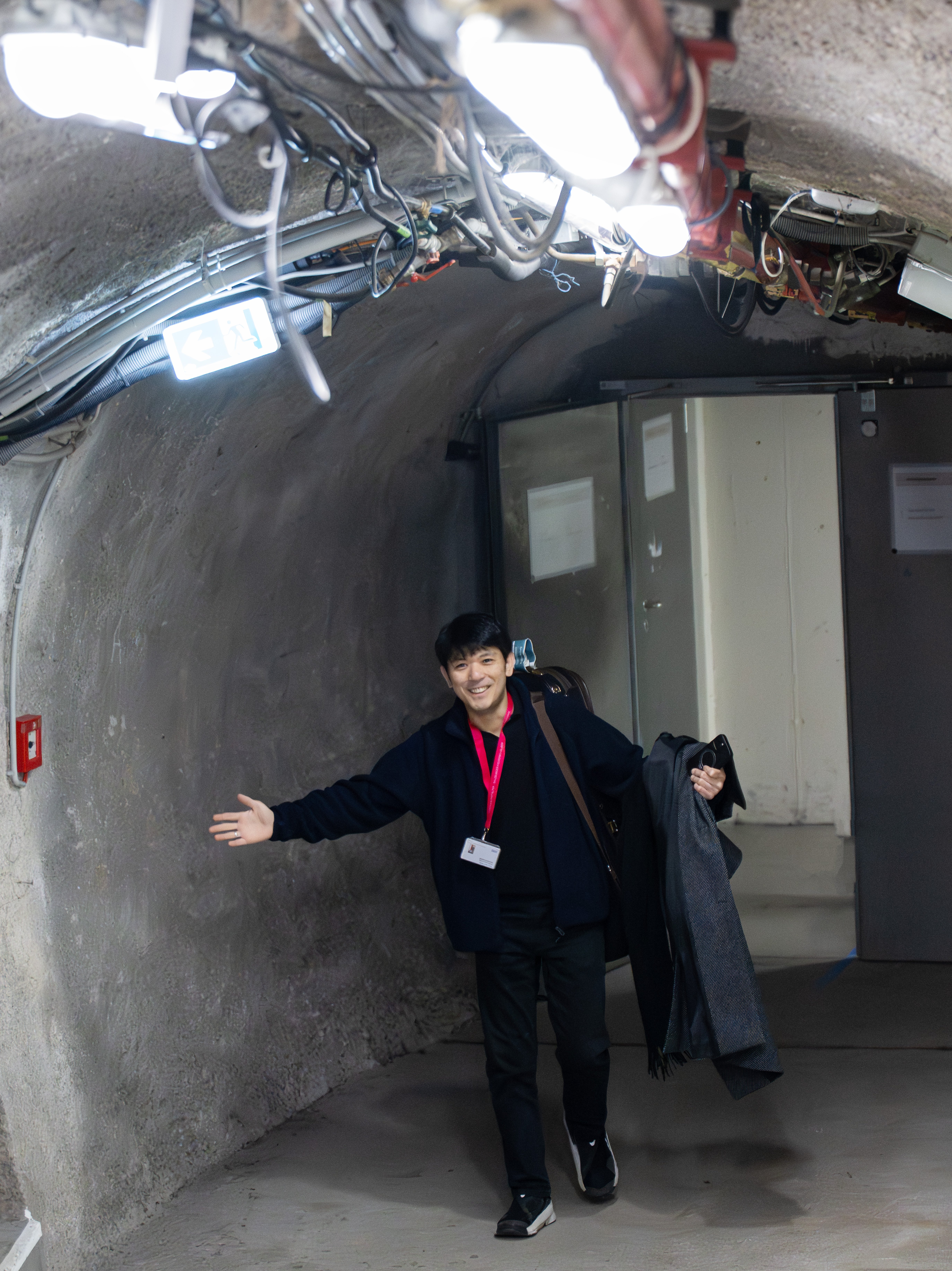 Eine Person steht in einem unterirdischen Tunnel mit freiliegenden Drähten, trägt einen Geigenkasten auf dem Rücken, hält einen Mantel und lächelt mit ausgebreiteten Armen.
