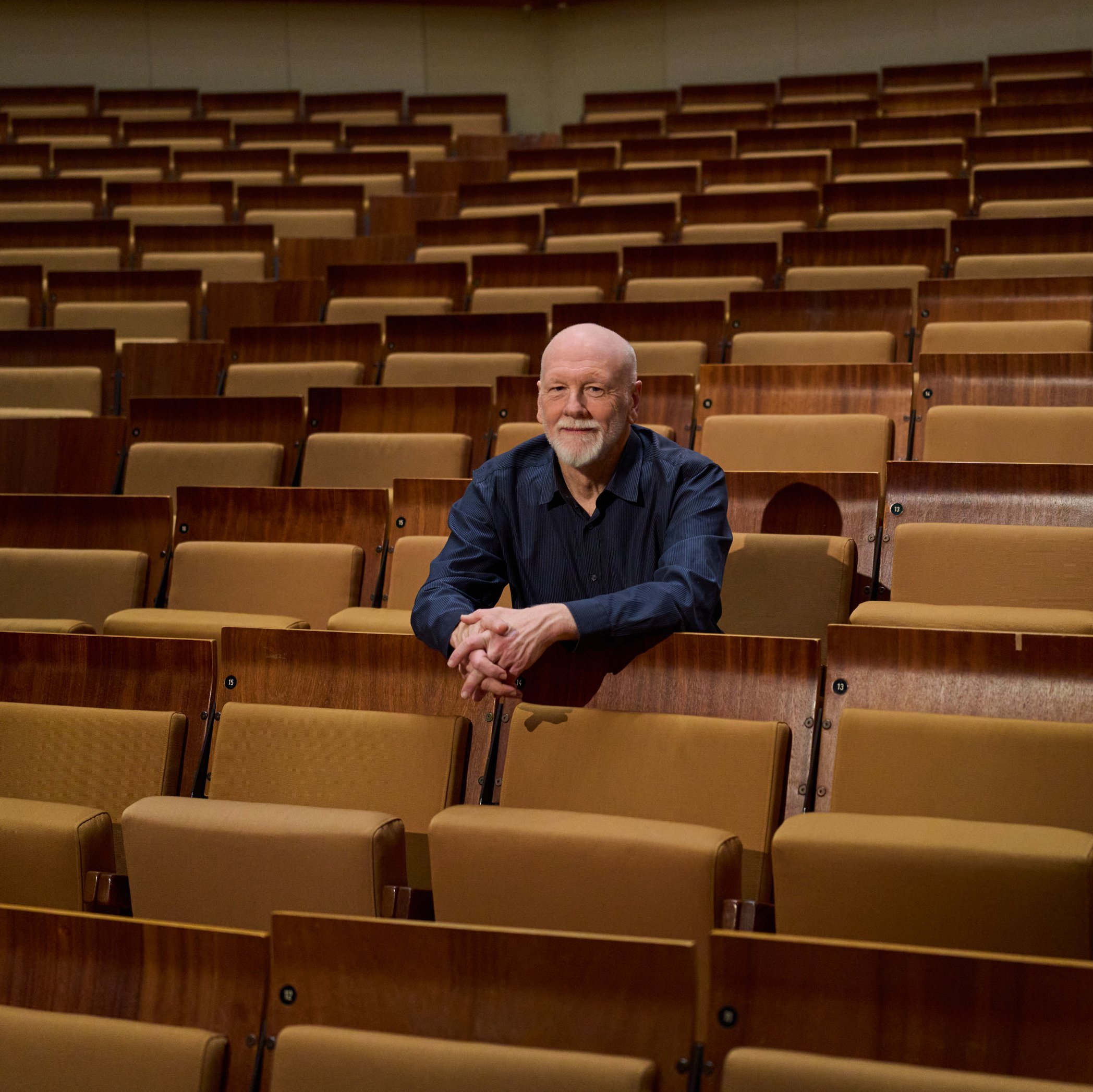 Brett Dean sitzt allein in der Mitte eines großen, leeren Saals mit braunen Sitzreihen und blickt nach vorne, die Arme auf den Sitz vor ihm gestützt.