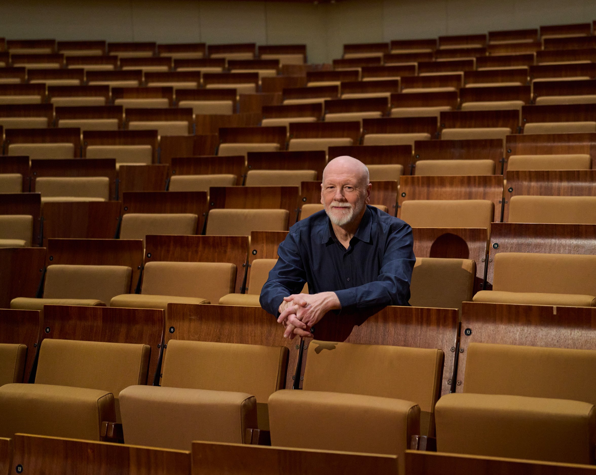 Brett Dean sitzt allein in der Mitte eines großen, leeren Saals mit braunen Sitzreihen und blickt nach vorne, die Arme auf den Sitz vor ihm gestützt.